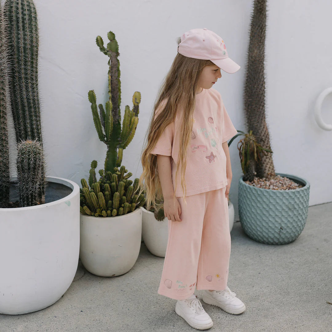 A young girl with long hair stands beside potted cacti, dressed in a pink outfit and cap, with her white sneakers visible beneath the Palm Beach Wide Leg Pants Terry Towel Palm Flamingo Pink by GOLDEN CHILDREN. The minimalistic and serene scene is enhanced by the backdrop featuring a white wall and a blue textured planter.