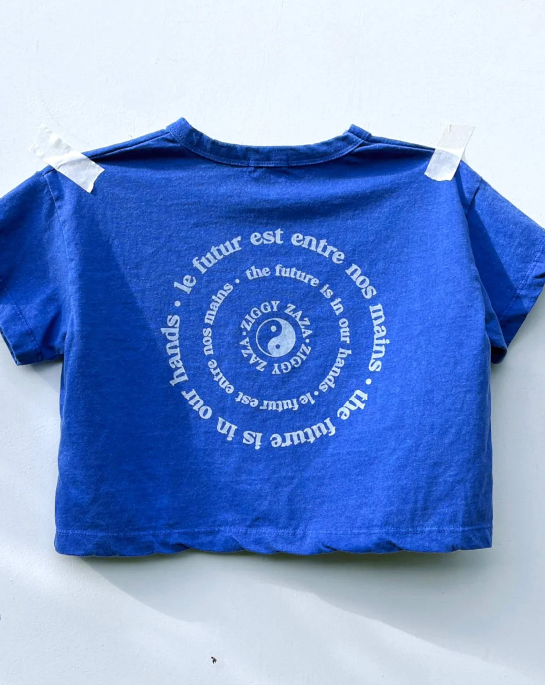 A relaxed, boxy fit Ziggy Zaza Love Your Mother Tee in Lapis Blue (Size 7-9) is taped to a wall. Crafted from premium lightweight washed cotton, it features a circular yin-yang design with the message "the future is in our hands" in French and English.