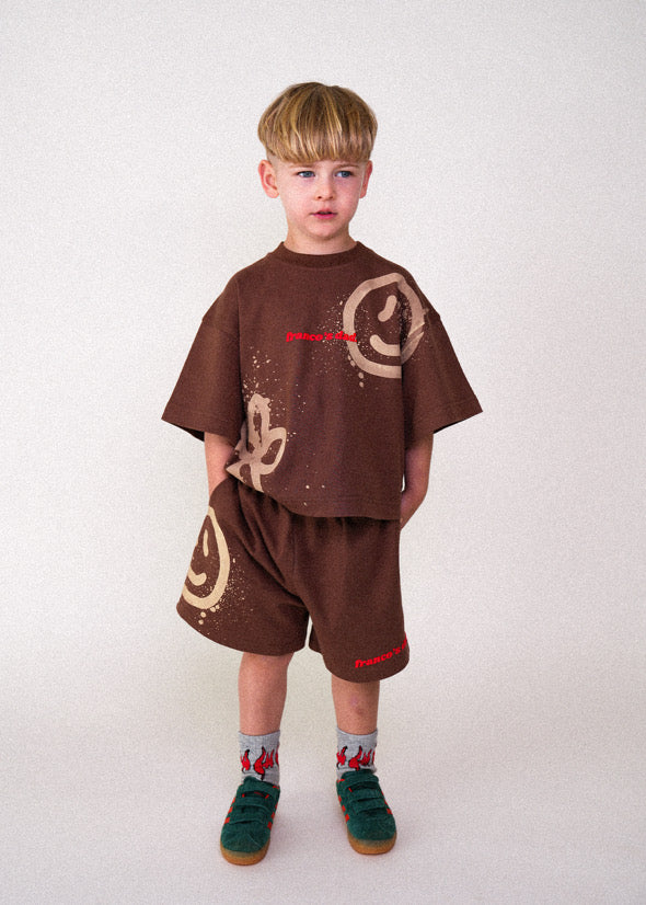 A young child wearing FRANCO'S DAD's brown Acid Dye Shorts in size 1 stands against a plain background. The oversized shirt with abstract designs and a small logo enhances their blonde hair, colorful socks, and green shoes perfectly.