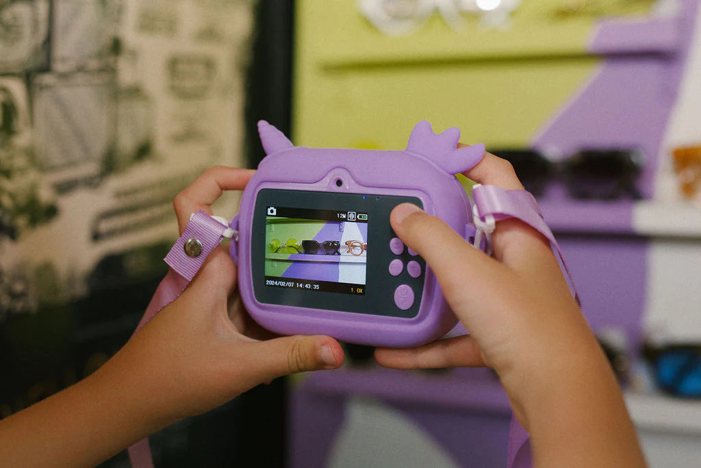 A child joyfully embraces creativity with the Oh My Frankie ~ Oh My Snap Instant Print Camera in purple, featuring cartoon ears. They capture an image of sunglasses against a vibrant background, delighting in the art of photography.