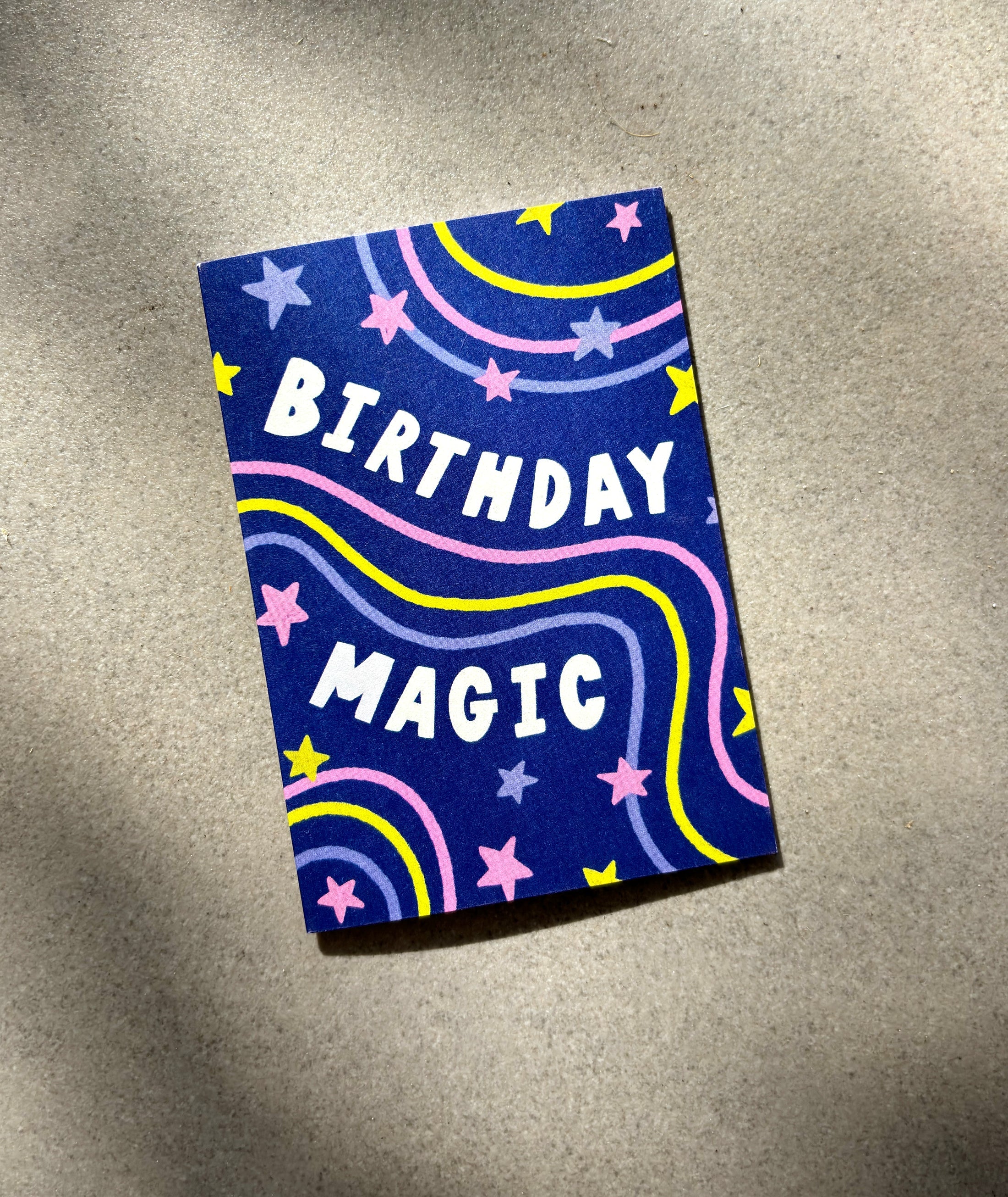 The Birthday Magic Mini Card by LAUREN SISSONS STUDIO has a dark blue background with the words "Birthday Magic" inscribed in white letters. Adorned with vibrant stars, moons, and swirling lines in shades of yellow, pink, and purple, this card is crafted from recycled paper and presented on a light brown surface.