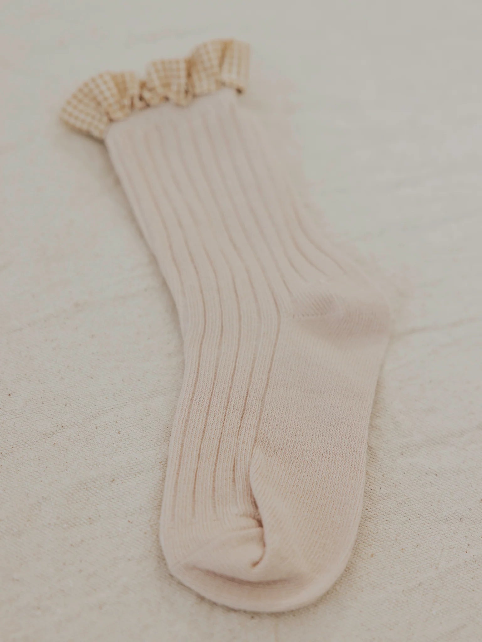 A single Frilly Socks Oat from VALENCIA BYRON BAY lies flat against a neutral fabric background. The light beige, ribbed sock features a ruffle trim in a gingham pattern at the top and possesses a subtle texture that looks soft and cozy.