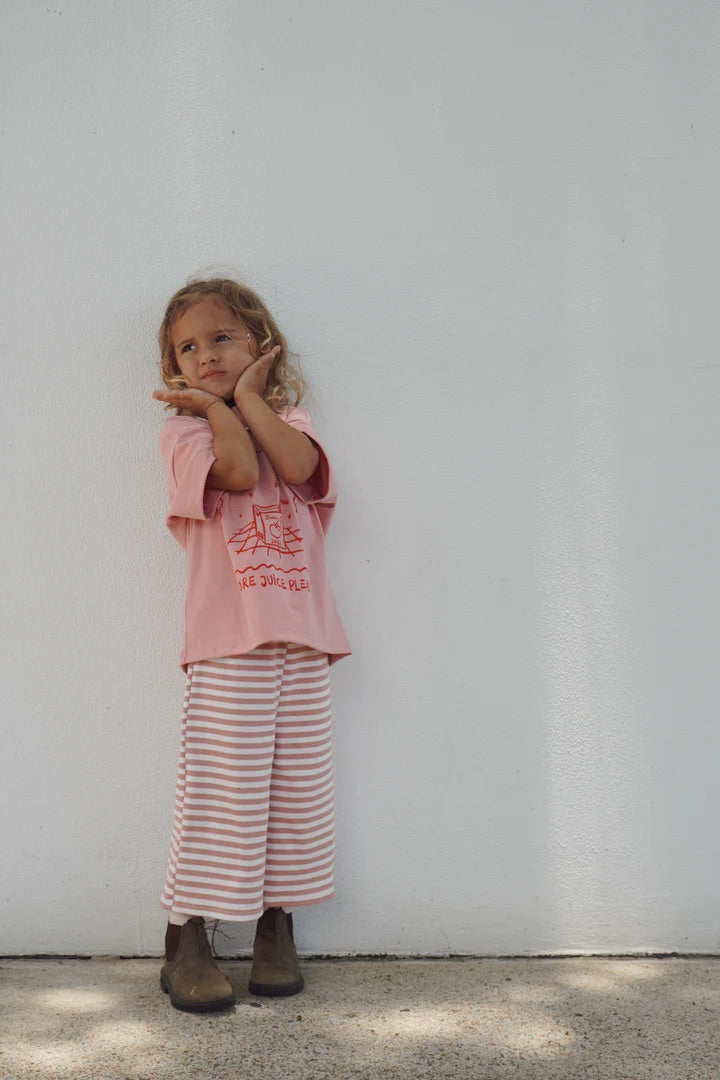 A young child with long curly hair stands against a white wall, wearing BOBBY G BABY WEAR's pink T-shirt made of organic cotton, Floaty Pants - Candy Cane Pink, and brown boots. They pose with hands on cheeks, looking thoughtful.