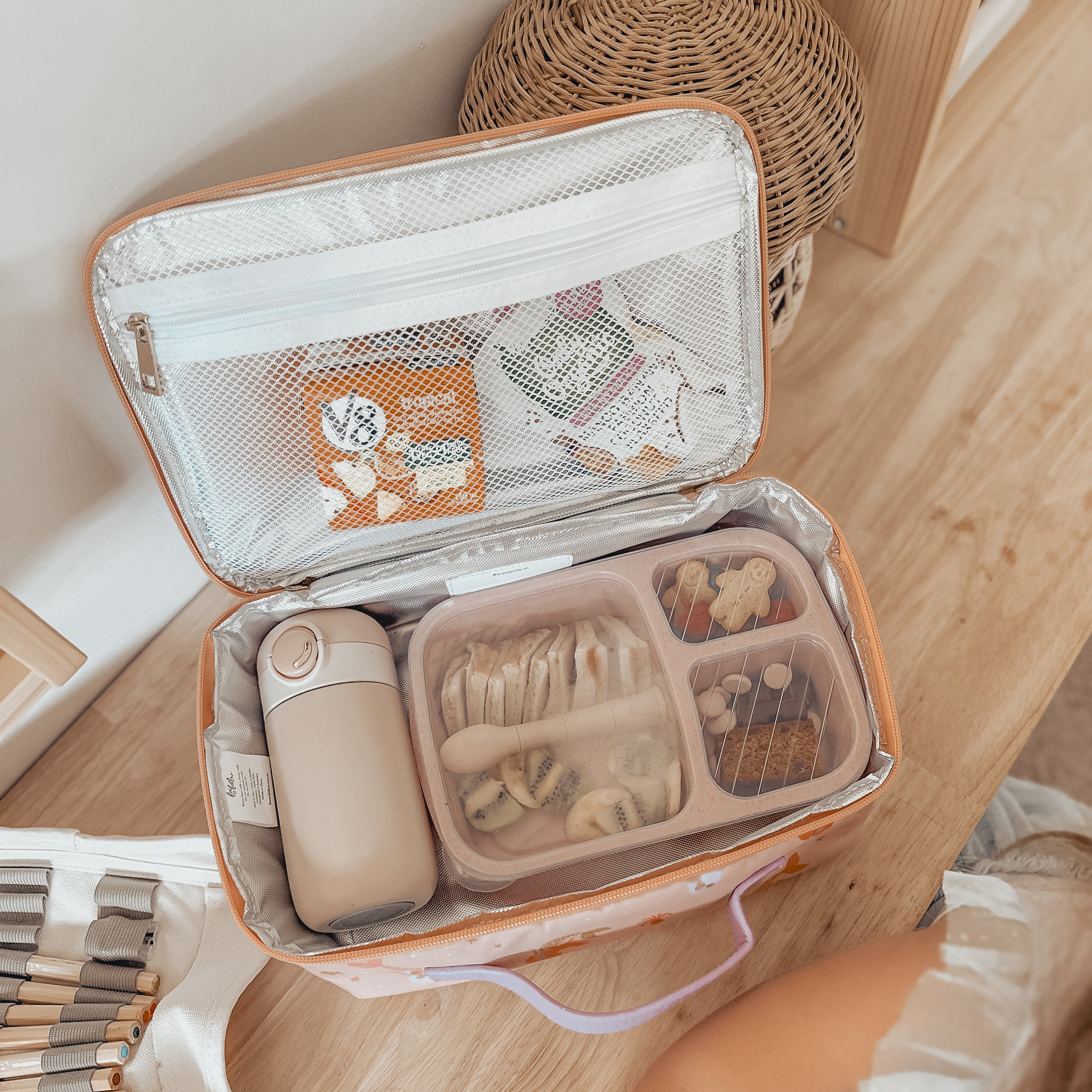 An open FOX & FALLOW Mermaids Lunch Bag on a wooden floor contains a beige thermos, a bento box filled with banana slices, nuts, and biscuits, and a zippered mesh pocket holding snacks. Made from waterproof fabric, this insulated lunch bag is set against the backdrop of a woven basket and a beige carpet.