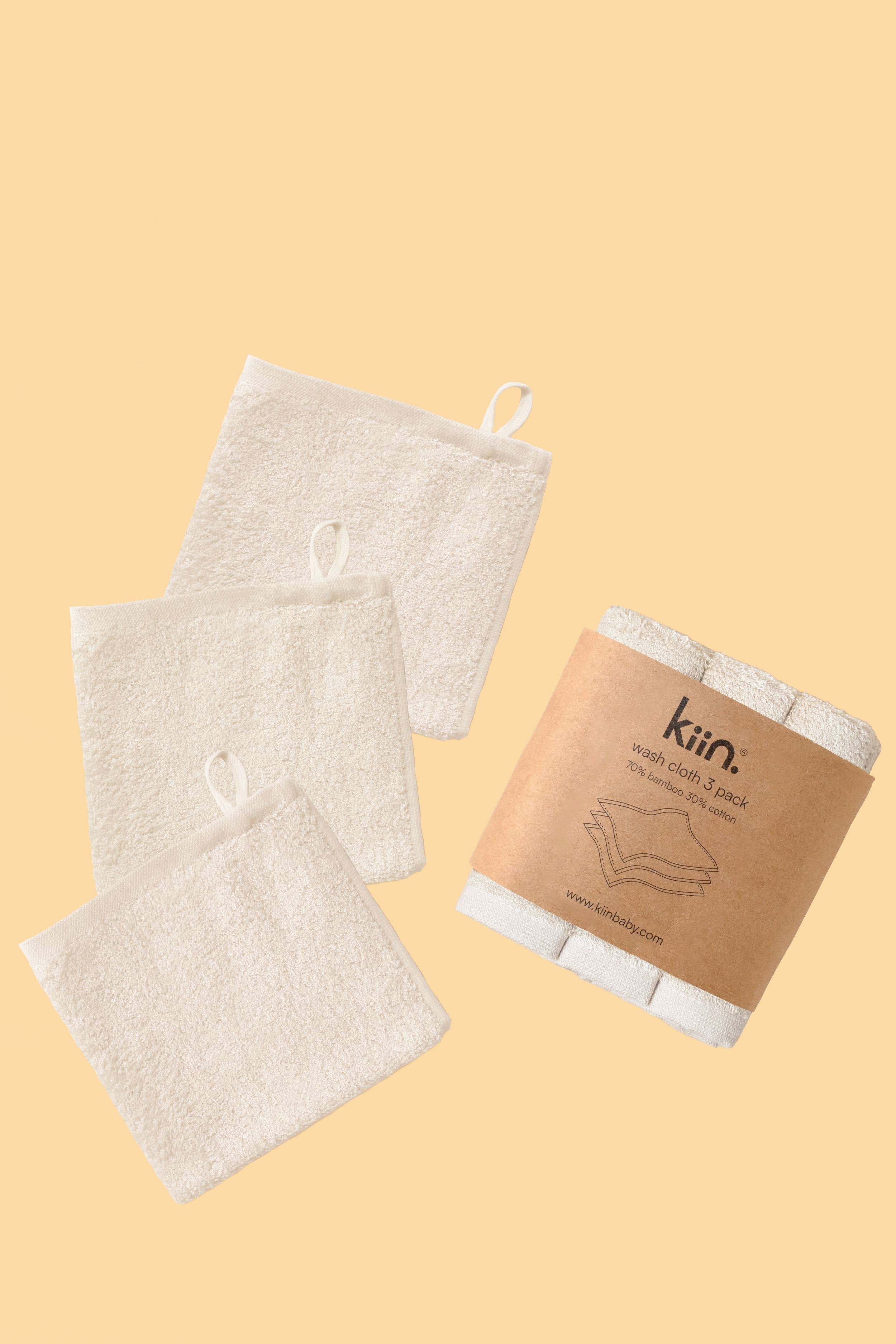 Three ivory KIIN BABY wash cloths—soft cotton-bamboo blend with hanging loops—are displayed flat, while a rolled five-pack in brown paper labeled "Kiin wash cloth 5 pack" sits alongside them on a light beige background.