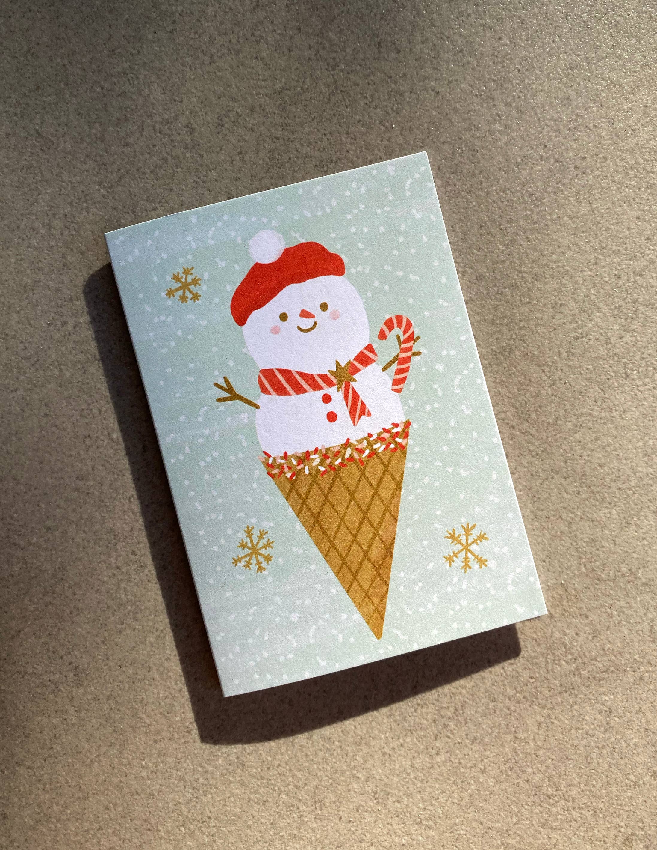 The Ms Snow Cone Mini Card from LAUREN SISSONS STUDIO features a cheerful snowman adorned with a red hat and scarf, holding a candy cane while nestled in an ice cream cone. Delicate snowflakes drift across the light green background of this charming illustrated card, which is thoughtfully printed on recycled paper for an eco-friendly appeal.