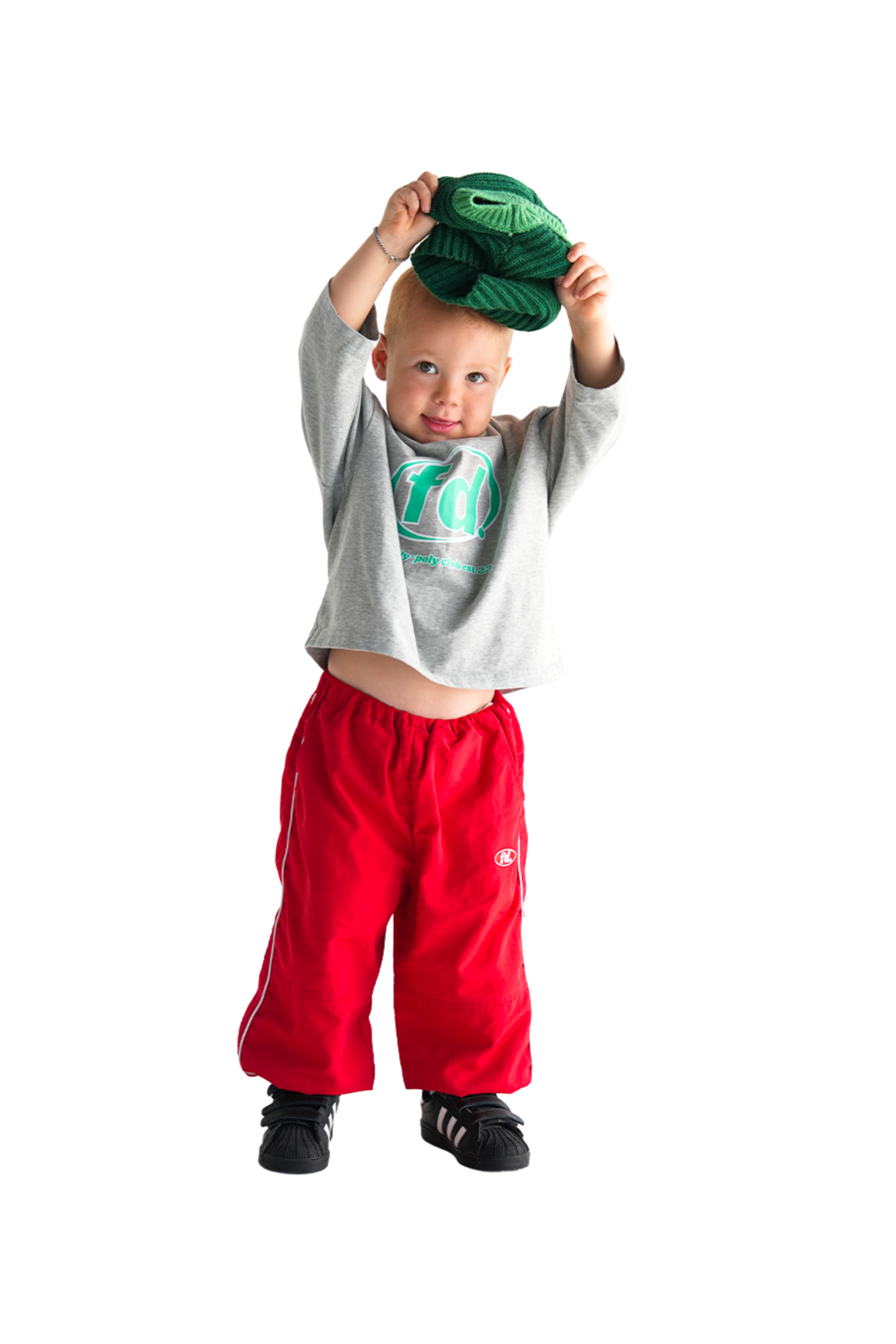 A cheerful child playfully lifts a green knit hat, wearing Franco's Dad Long Sleeve Tee Grey Marle by FRANCO'S DAD with red pants and black shoes against a white background.