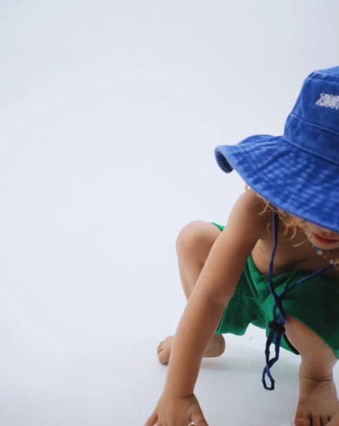 A child crouches in a green outfit wearing the Ziggy Zaza ~ Everyday Scout Hat Blu by ZIGGY ZAZA, set against a plain white background. The adjustable chin strap and cotton canvas brim partially hide their face.