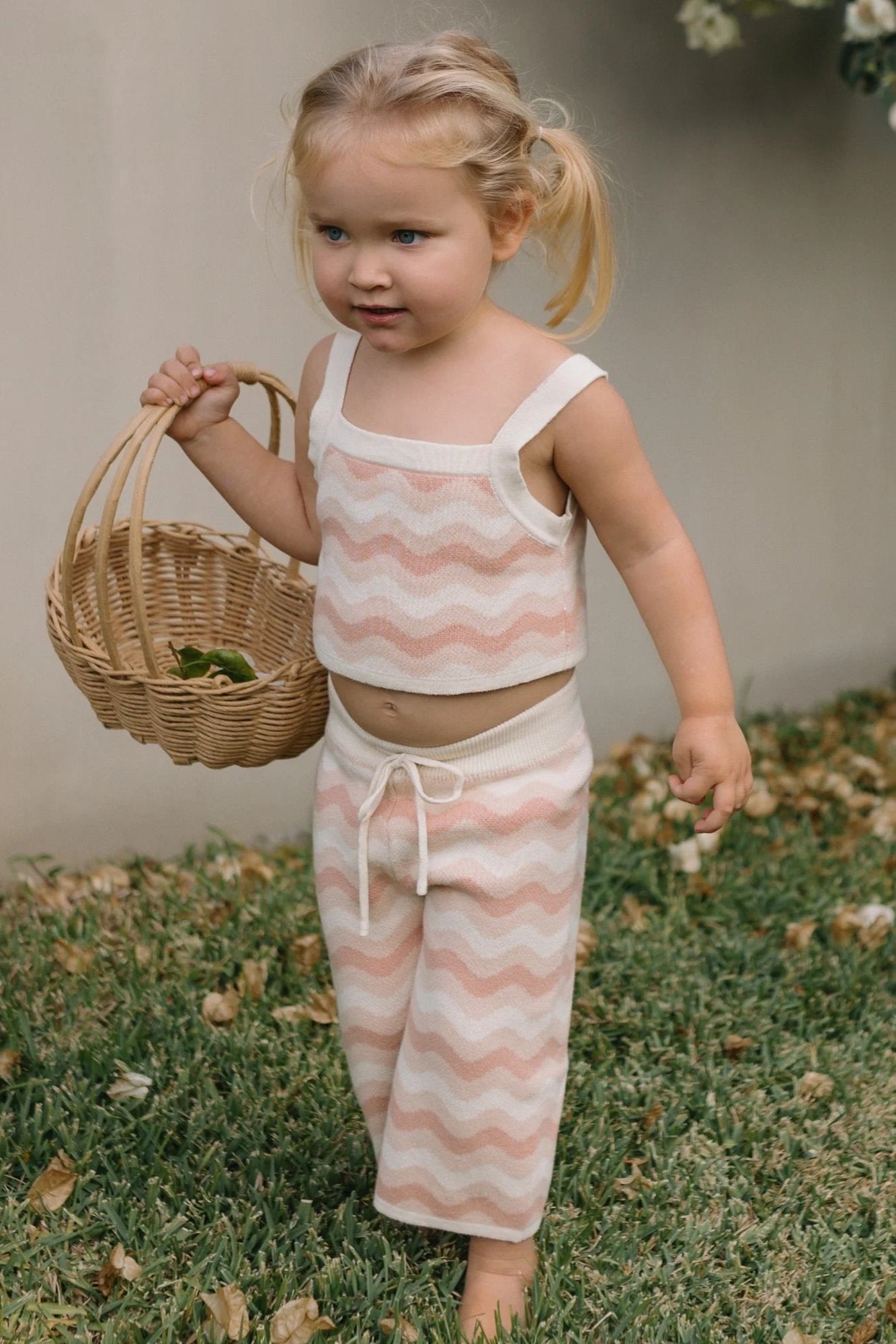 A young girl with blonde pigtails walks on the grass, holding a wicker basket. She's wearing the Seashore Knit Set Strawberry Cream from GOLDEN CHILDREN, featuring a seaside-inspired design in relaxed fit cotton with pink and white zigzag patterns. Dry leaves are scattered around her feet.