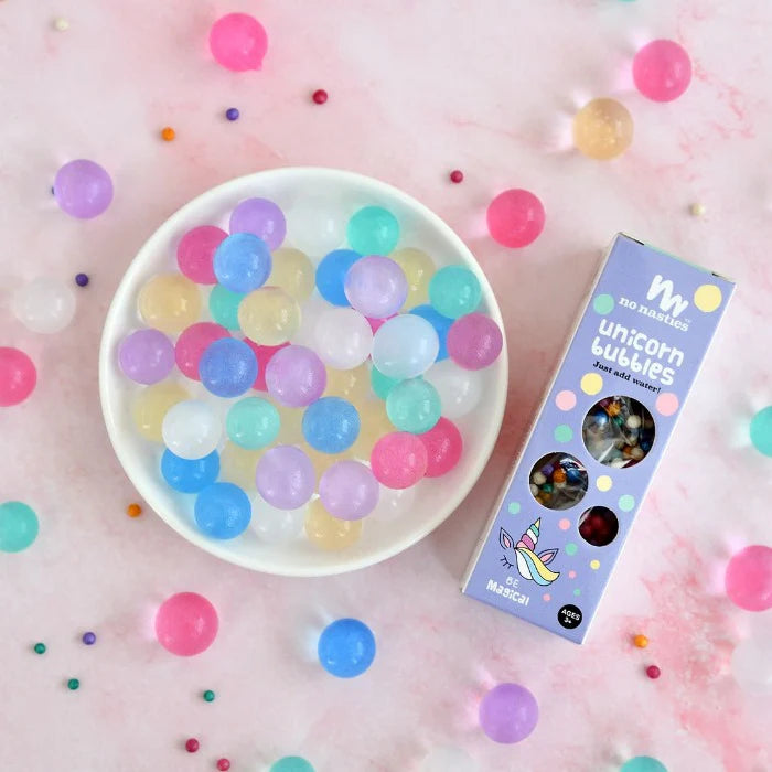 A white bowl contains vibrant, translucent Unicorn Bubbles Pastel Biodegradable Water Beads from NO NASTIES, beautifully displayed on a pink marble surface for sensory play. Small, multicolored beads are scattered around to enhance fine motor skill development. A box labeled "Unicorn Bubbles" sits on the right.