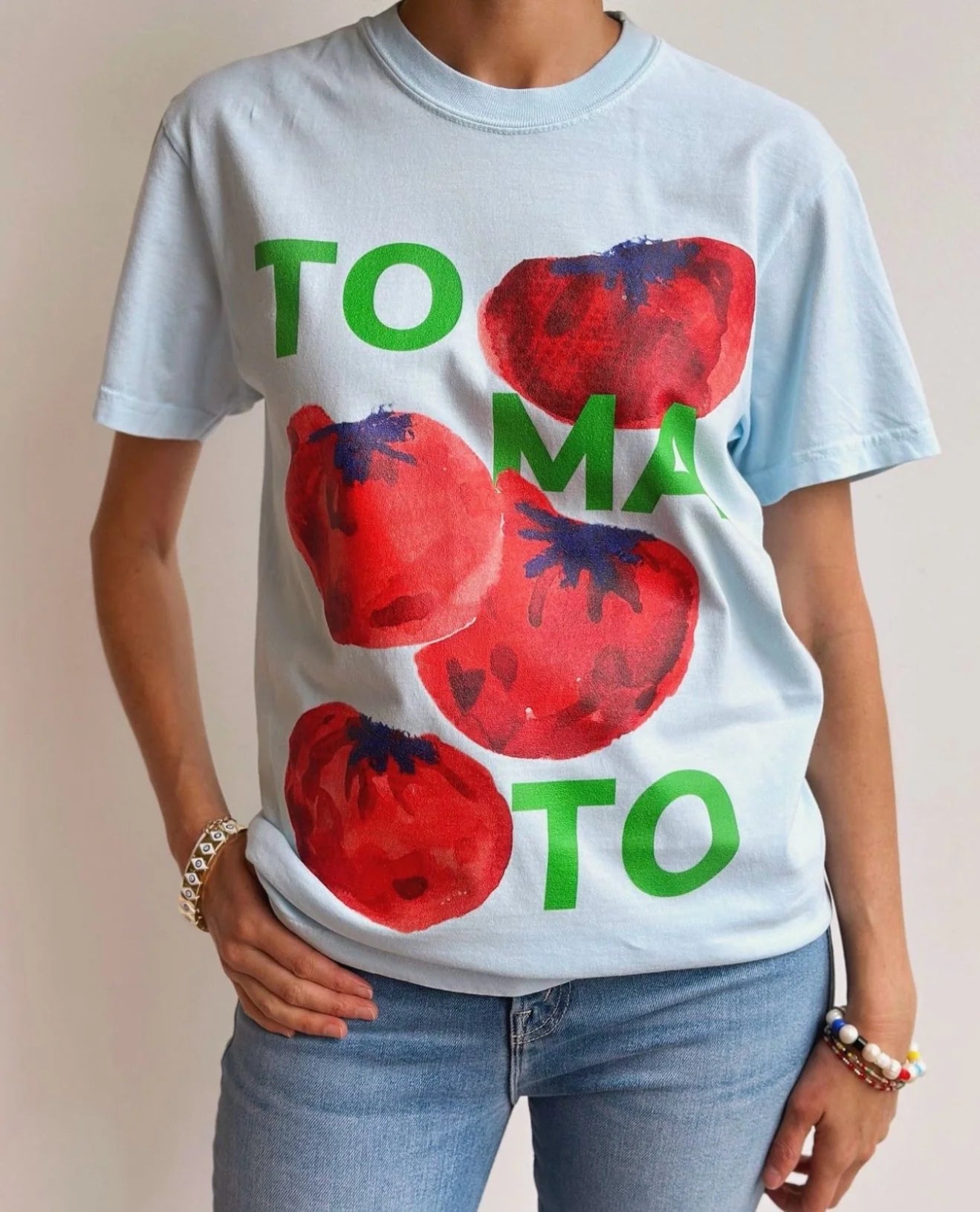 A person models a MAKU THE LABEL Tomato T-Shirt, featuring a light blue background with large red tomatoes and "TOMATO" in green. Offering a classic fit, they're styled with jeans and colorful bracelets, posing confidently against a plain white backdrop.