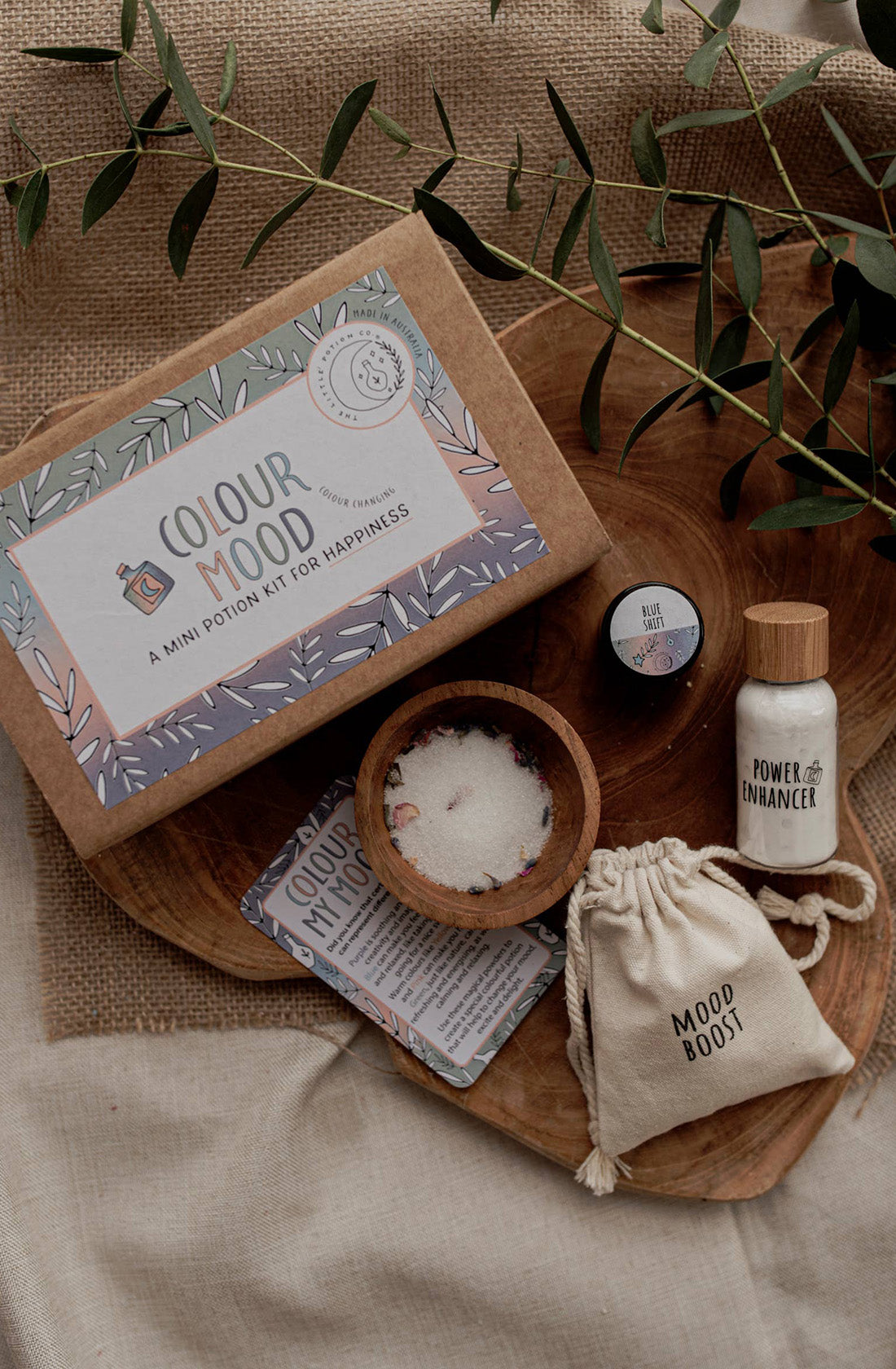 A tabletop with THE LITTLE POTION CO's MINI Colour Mood Potion Kit, featuring a brown box with floral designs, a small wooden bowl with white mixture, a "Mood Boost" drawstring pouch, a "Power Enhancer" bottle, a small container, a card, and eucalyptus leaves scattered around—perfect for sensory play.