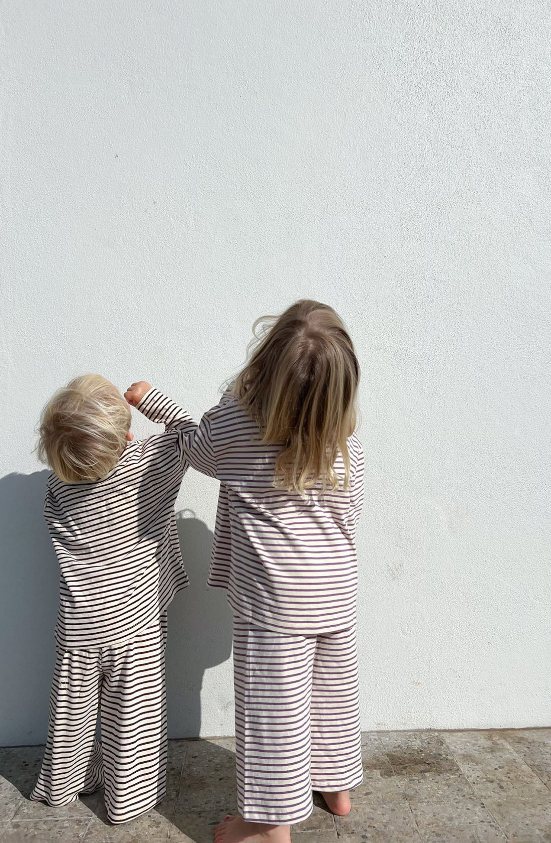 Two small children with blonde hair stand barefoot against a plain white wall. They are wearing matching pajamas from LENN LABEL’s Every Day Set Lavender, made of 100% knit cotton. One child reaches up toward the wall while the other gazes upward. The elastic waistband adds comfort, and the scene is brightly lit, suggesting daylight.