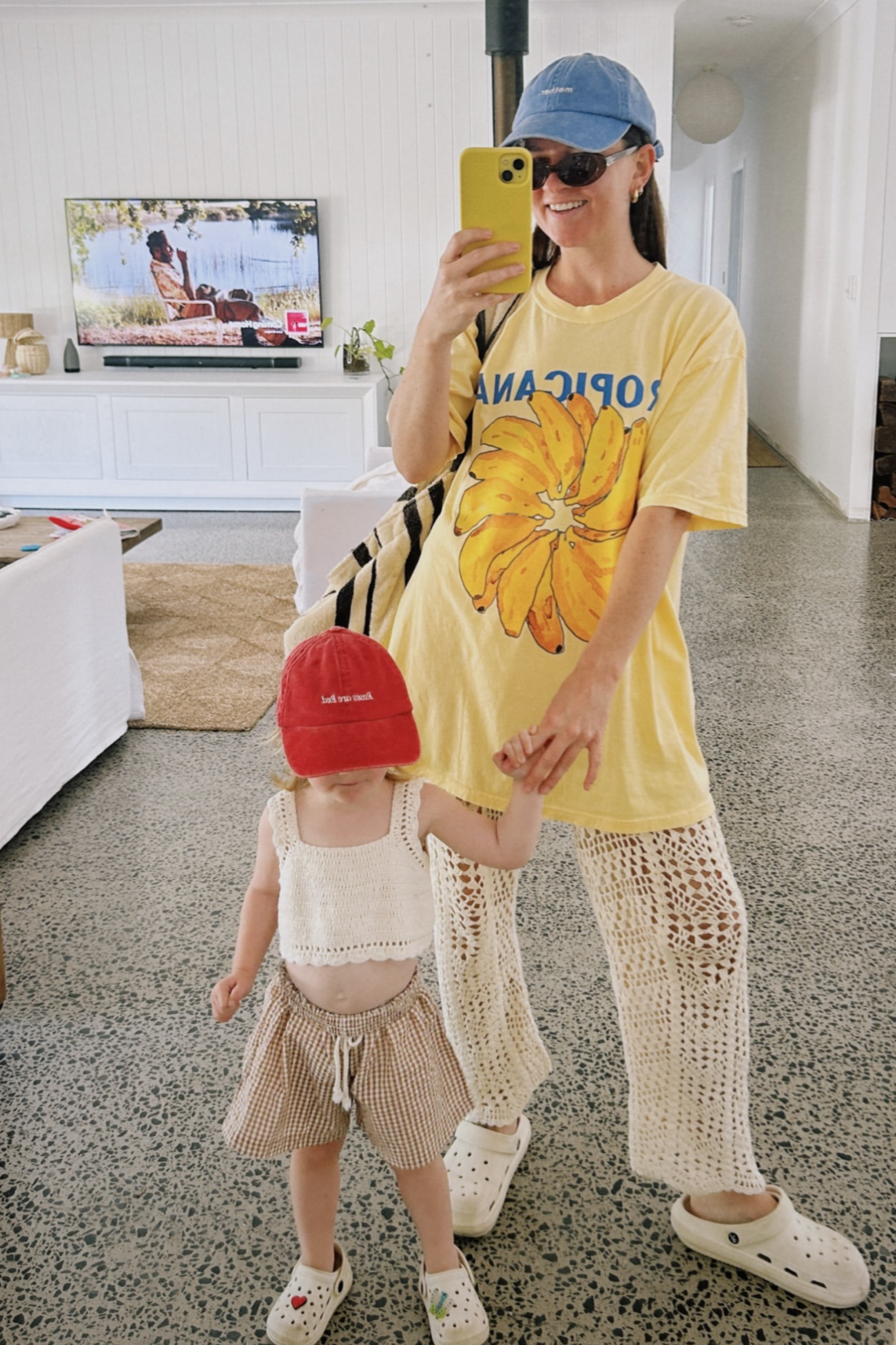 A woman wearing a yellow, hand-painted Tropicana Banana T-shirt by MAKU FENAROLI and crochet pants takes a mirror selfie with a child dressed in a red cap, crochet top, and striped shorts. Both are sporting Crocs. In the background of the lightly decorated room, the TV is playing, featuring unisex tee styles crafted from 100% cotton.