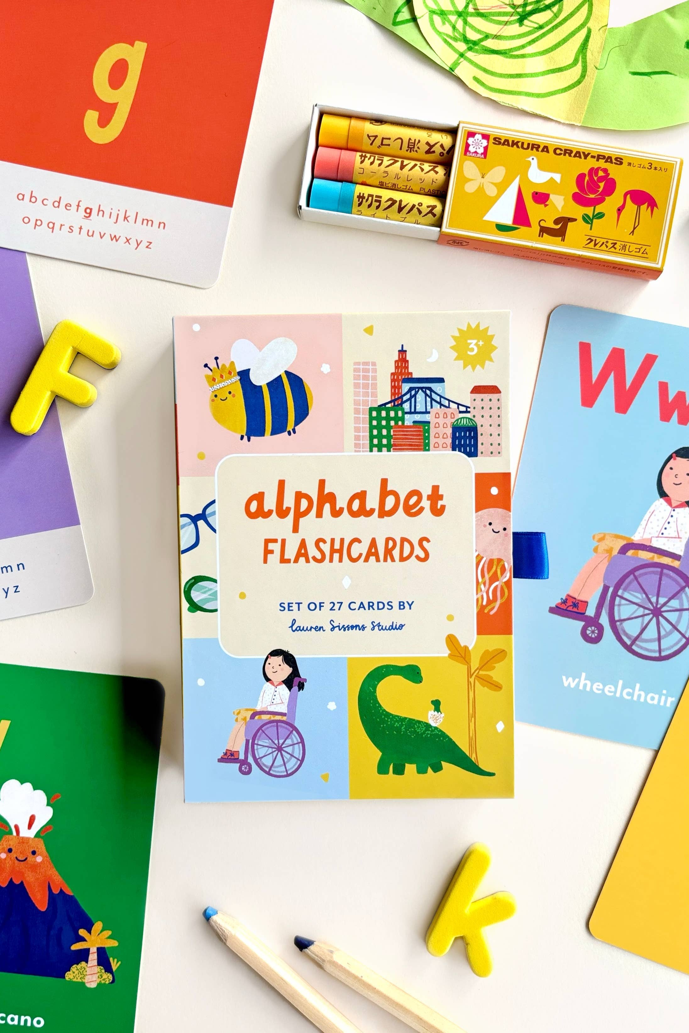 Image showcasing the "A-Z Illustrated Alphabet Flashcards" in vibrant packaging by LAUREN SISSONS STUDIO. The featured card displays illustrations of a bee, a child in a wheelchair, and city buildings. Encircling the set are additional laminated flashcards, crayons, alphabet letters, and pencils—ideal educational toys for children.