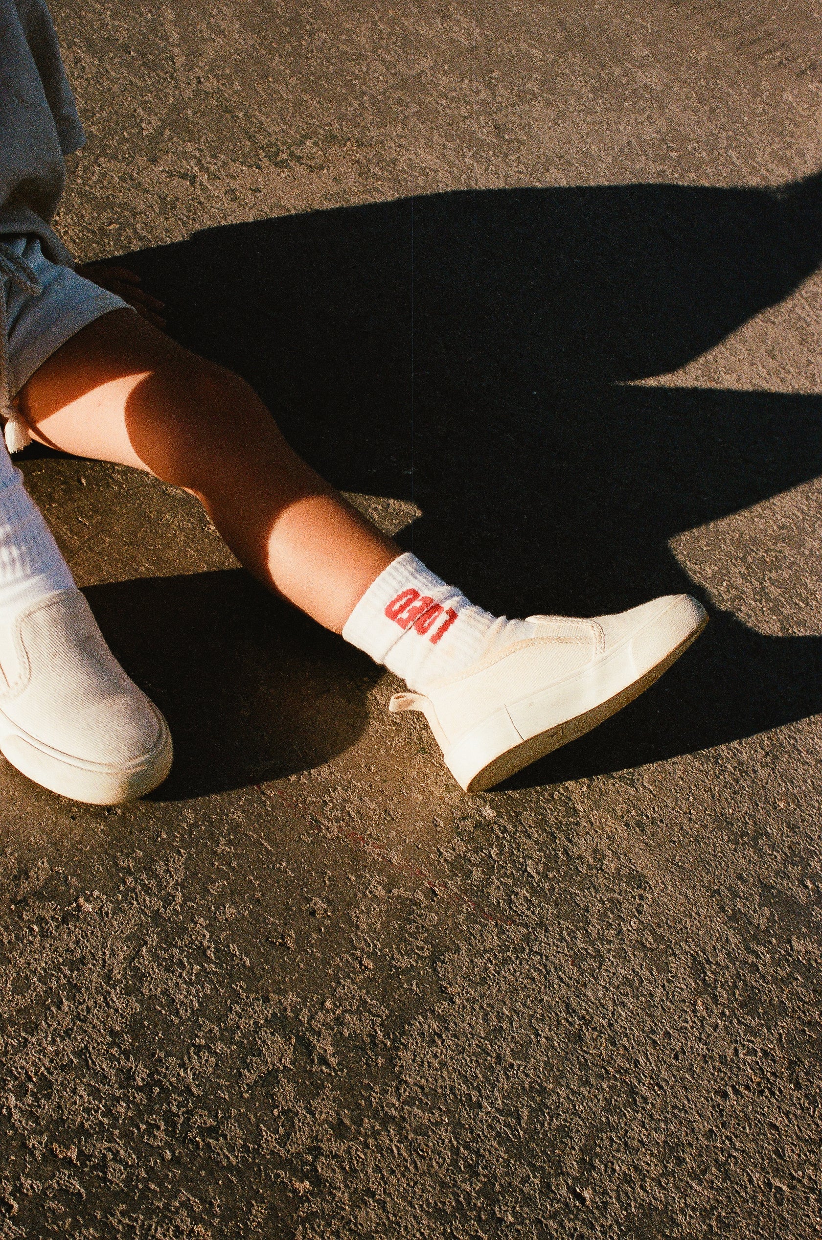 A person is sitting on a concrete surface with one leg extended. They are wearing white slip-on shoes, light-colored shorts, and white sports socks from TINY LOVE CLUB featuring the "Loved Sock" design that says "GOLD" in red letters. The image captures a shadow on the ground beside their leg.