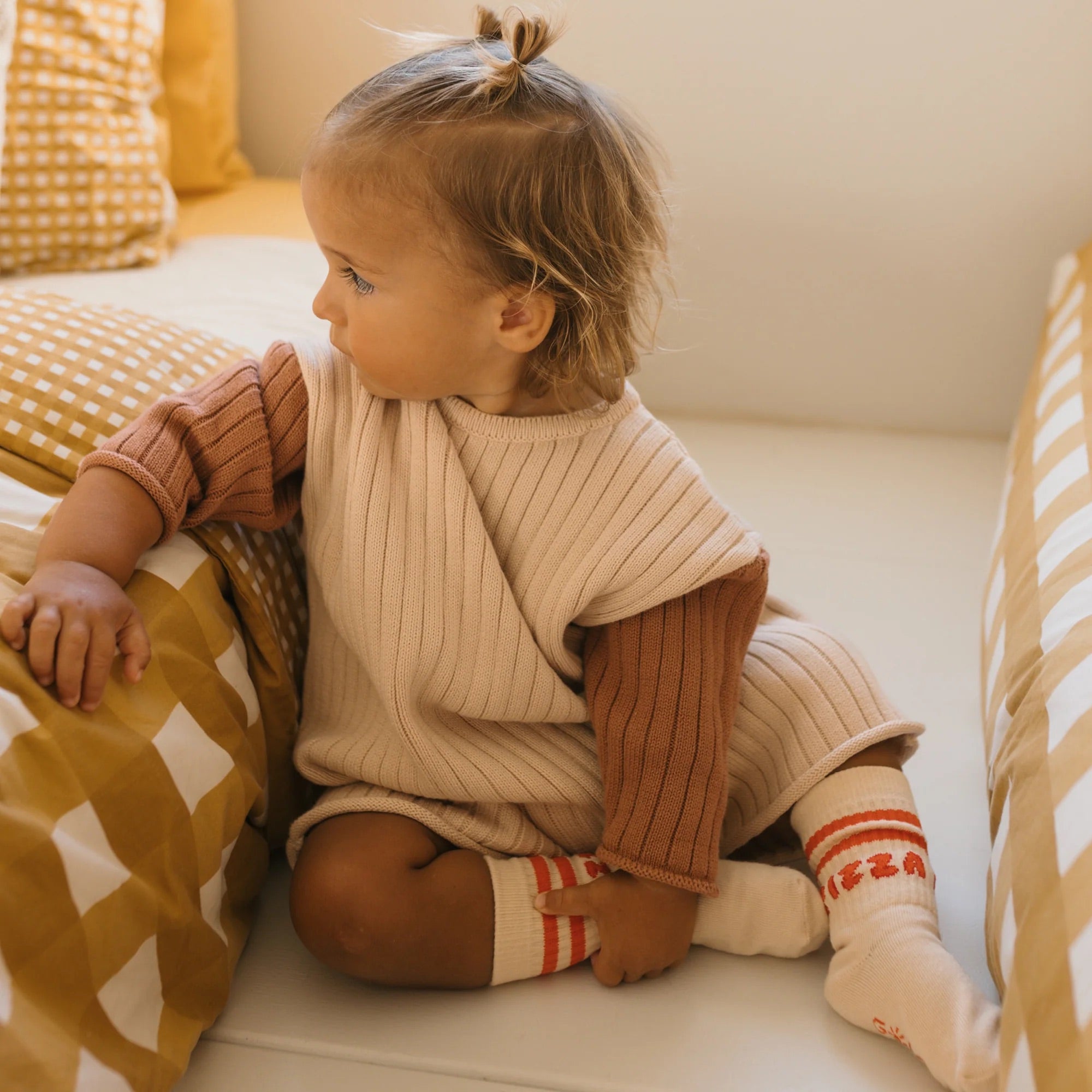 A toddler with light brown hair in a small ponytail sits on a bed. The child is wearing the "Golden Knit Romper Caramel Sundae" by GOLDEN CHILDREN, featuring brown sleeves and paired with cream socks adorned with orange stripes and red text. The bed, adorned with white and mustard checkered bedding, enhances the cozy feel of the neutral beige yarn attire.