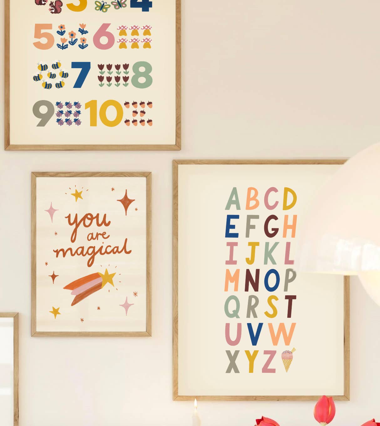 Three framed artwork pieces adorn the nursery wall. From left: an educational number chart from 1 to 10, a "You are magical" print with stars and a rainbow, and the Bold A-Z Alphabet Wall Art Print by LAUREN SISSONS STUDIO with letters in different colors. A lamp and red flowers are partially visible in the foreground.