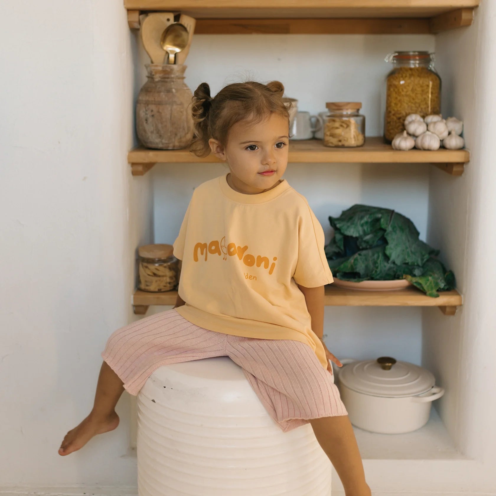 A young child with light brown hair styled in two pigtails sits on a large white container in a kitchen. The child is wearing a GOLDEN CHILDREN Macaroni Tee Golden Butter made from soft stretch fabric and light pink pants. Behind them, wooden shelves display jars of food, garlic bulbs, and leafy greens.