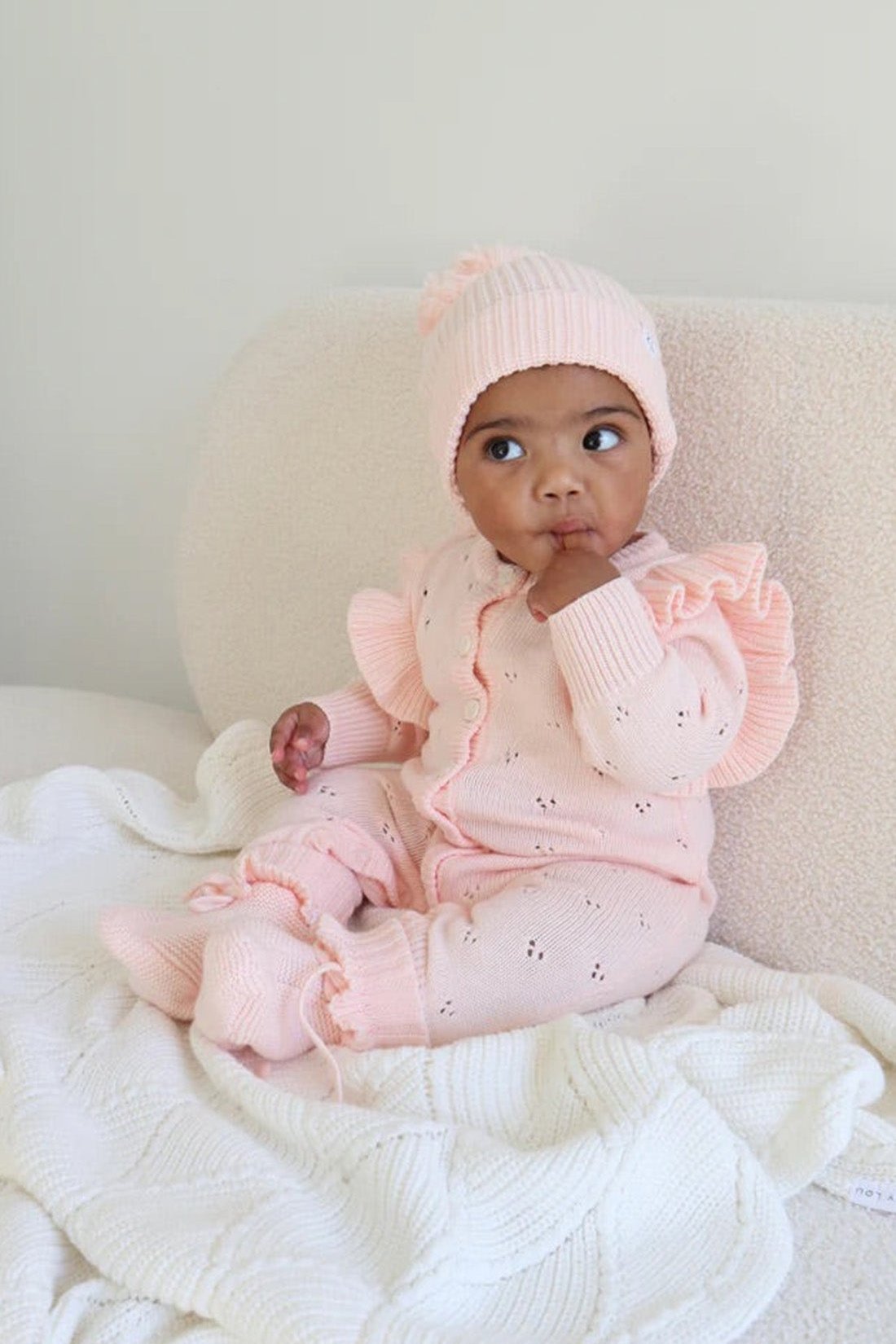 A baby wearing a pink knit outfit with ruffles and a matching ZIGGY LOU Beanie Primrose sits on a soft, off-white blanket. The baby looks to the side with a curious expression, and has one hand near their mouth.