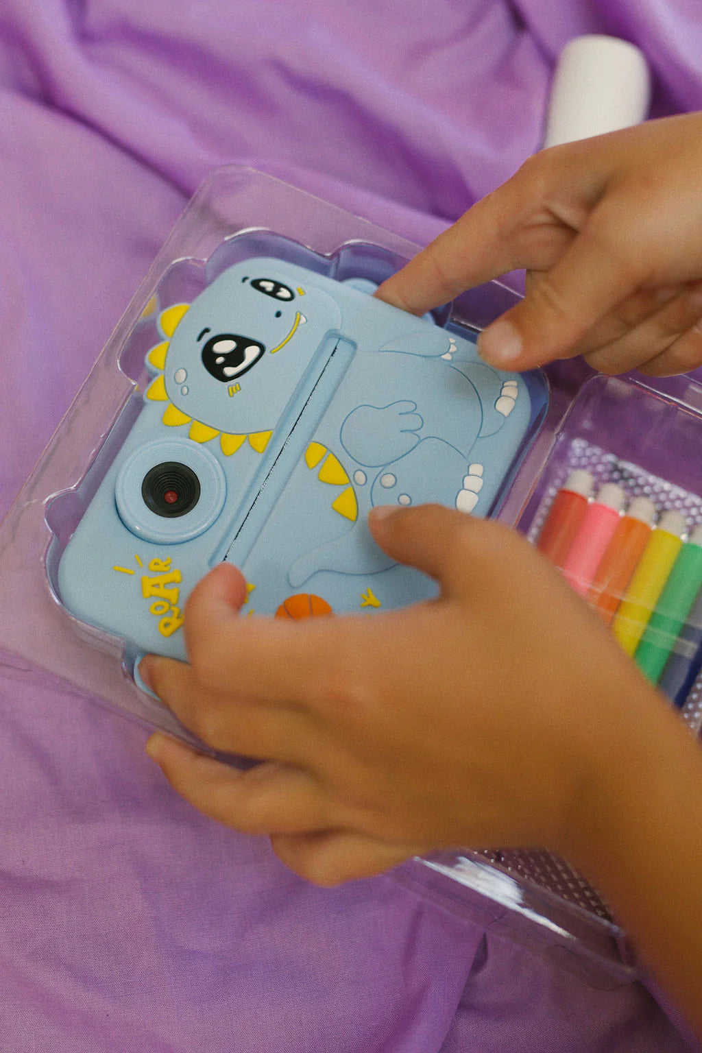 Hands holding the blue "Oh My Snap Instant Print Camera" by OH MY FRANKIE, featuring playful yellow and black accents. Ideal for creative play, it sits beside colorful markers on a soft purple fabric background.