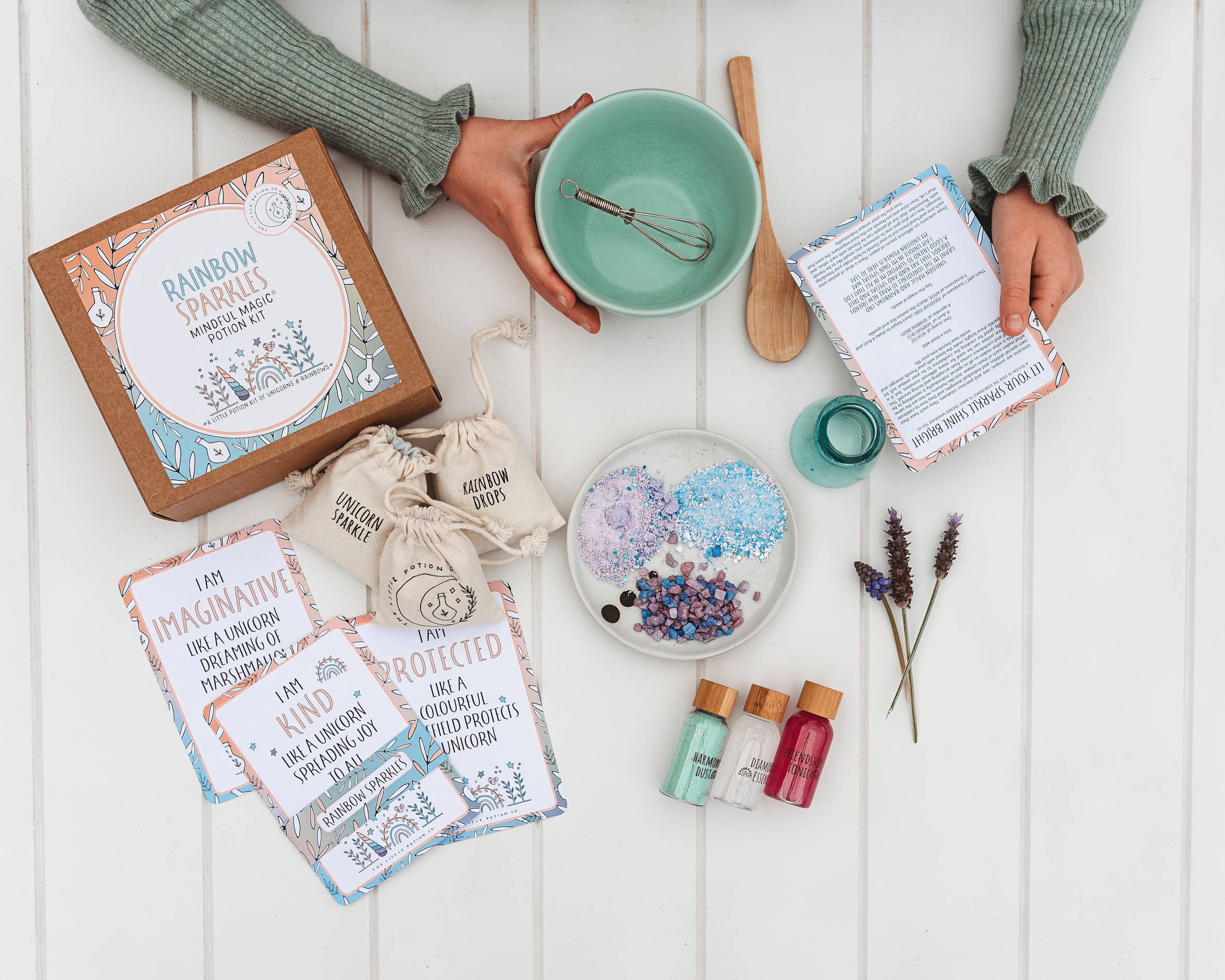 Top-down view of a table set for a DIY craft project. Includes THE LITTLE POTION CO's "Rainbow Sparkles MINDFUL Potion Kit," instruction cards, a whisk, mixing bowl, small bottles of colorant, and dried lavender flowers. Perfect for sensory play or even as magic potion kits. Two hands are holding instruction papers amidst beads and craft materials.