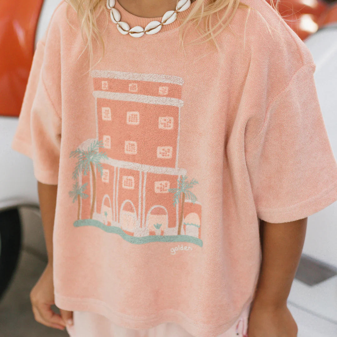 A person sporting a peach-hued Room Service Mid Sleeve Tee Terry Towel Spanish Villa by GOLDEN CHILDREN, featuring a graphic of the Colony Hotel and palm trees. They're also wearing a shell necklace while standing in front of sections of a red and white vehicle.