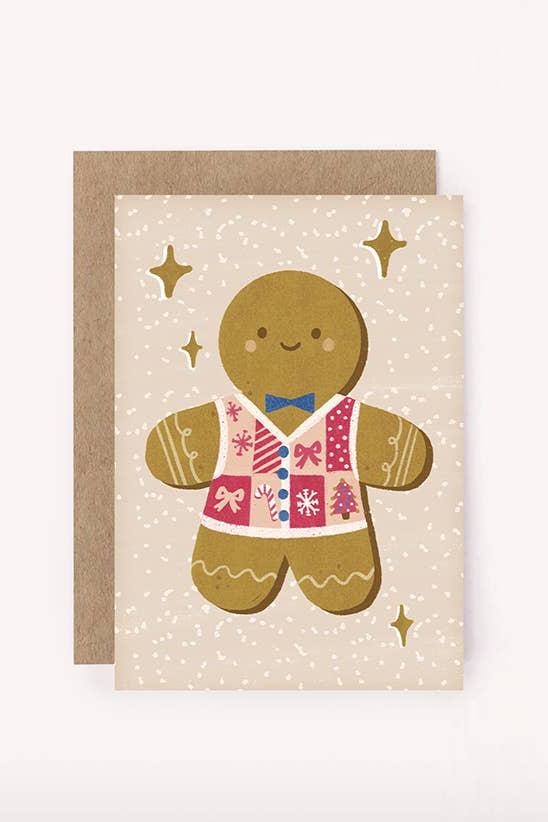 The Gingerbread Mini Card by LAUREN SISSONS STUDIO features a cheerful gingerbread man wearing a vibrant Christmas sweater, adorned with a blue bowtie and a smile. It is set against a beige background decorated with white speckles and golden star accents, and comes paired with an eco-friendly brown recycled paper envelope.