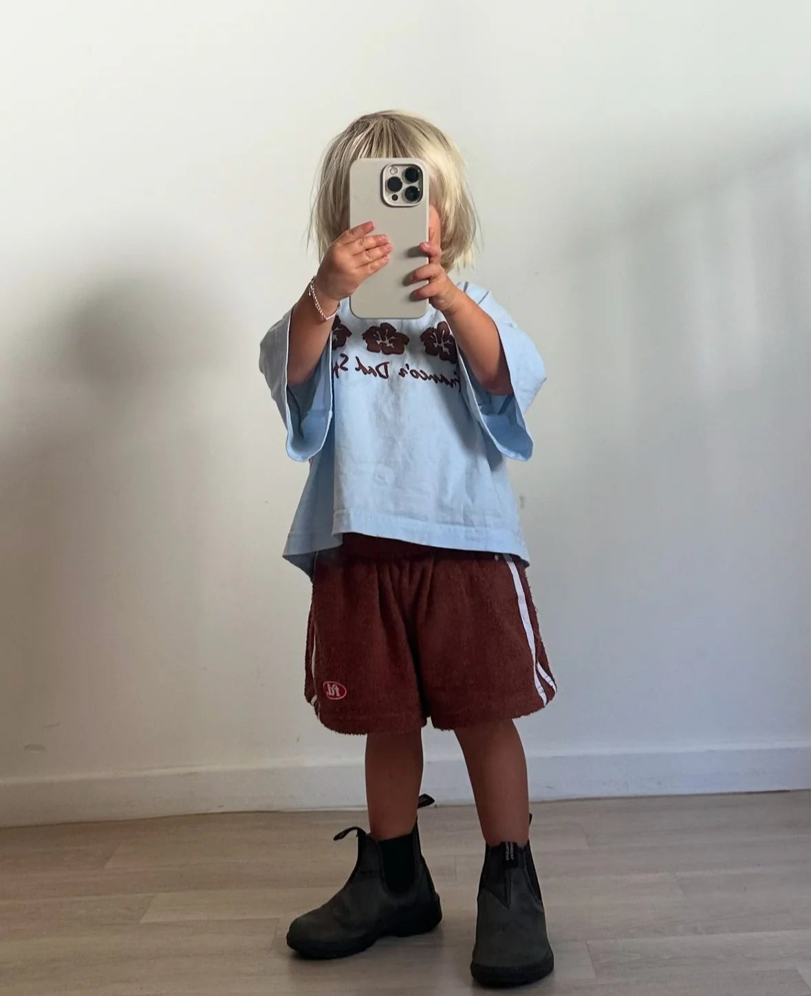 A child, wearing an oversized light blue PRE-ORDER Hibiscus Tee by FRANCO'S DAD, with tousled blonde hair, captures a snapshot on a smartphone. They pair the tee with red shorts and black boots while standing against a plain white wall on a wooden floor.