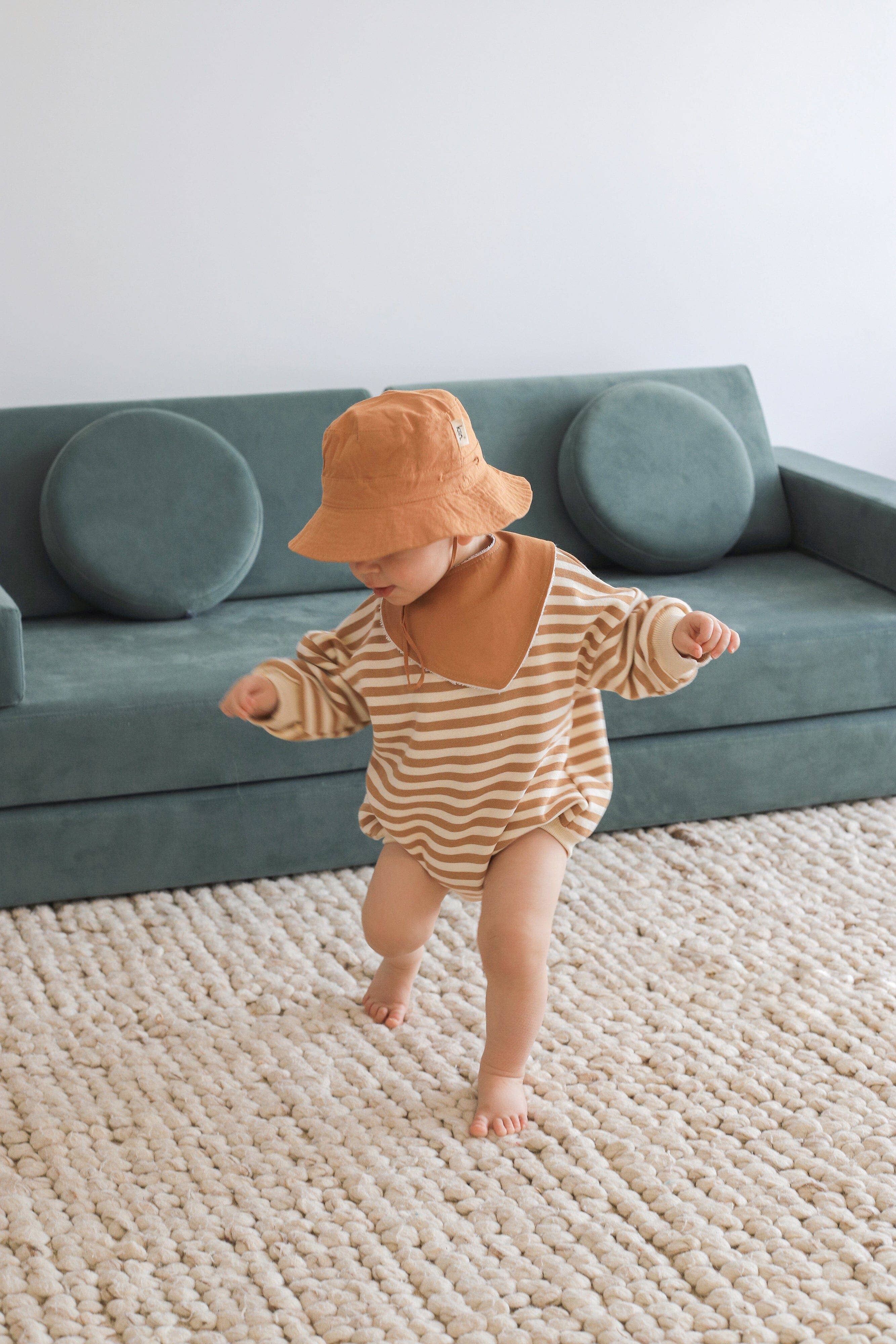 A toddler in a striped outfit and hat explores an absorbent bamboo/cotton carpet, with a green sofa and circular cushions nearby. A Kiin Baby Dribble Bib is playfully tossed aside, creating a playful and cozy atmosphere.