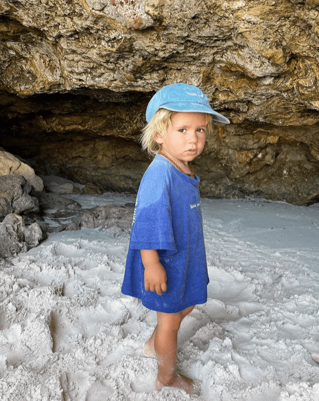 A young child, clad in a Ziggy Zaza Love Your Mother Tee Lapis Blue (Size 7-9) by ZIGGY ZAZA, is barefoot on sandy ground before a rocky cave. Blonde hair peeks from under a blue cap as natural light softly illuminates the scene.