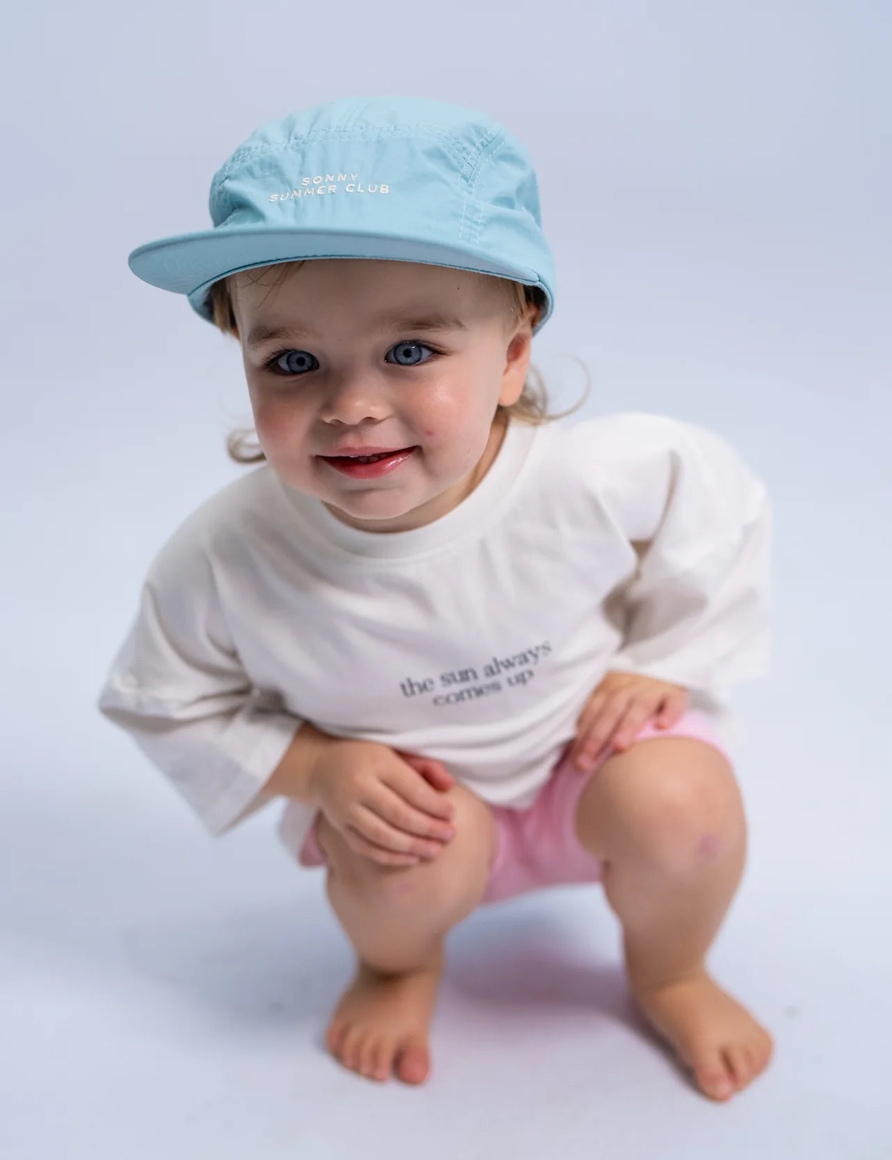 A young child wearing the Summer Club Cap : Ocean by SONNY LABEL, along with a white shirt and pink shorts, crouches and smiles at the camera against a plain background.