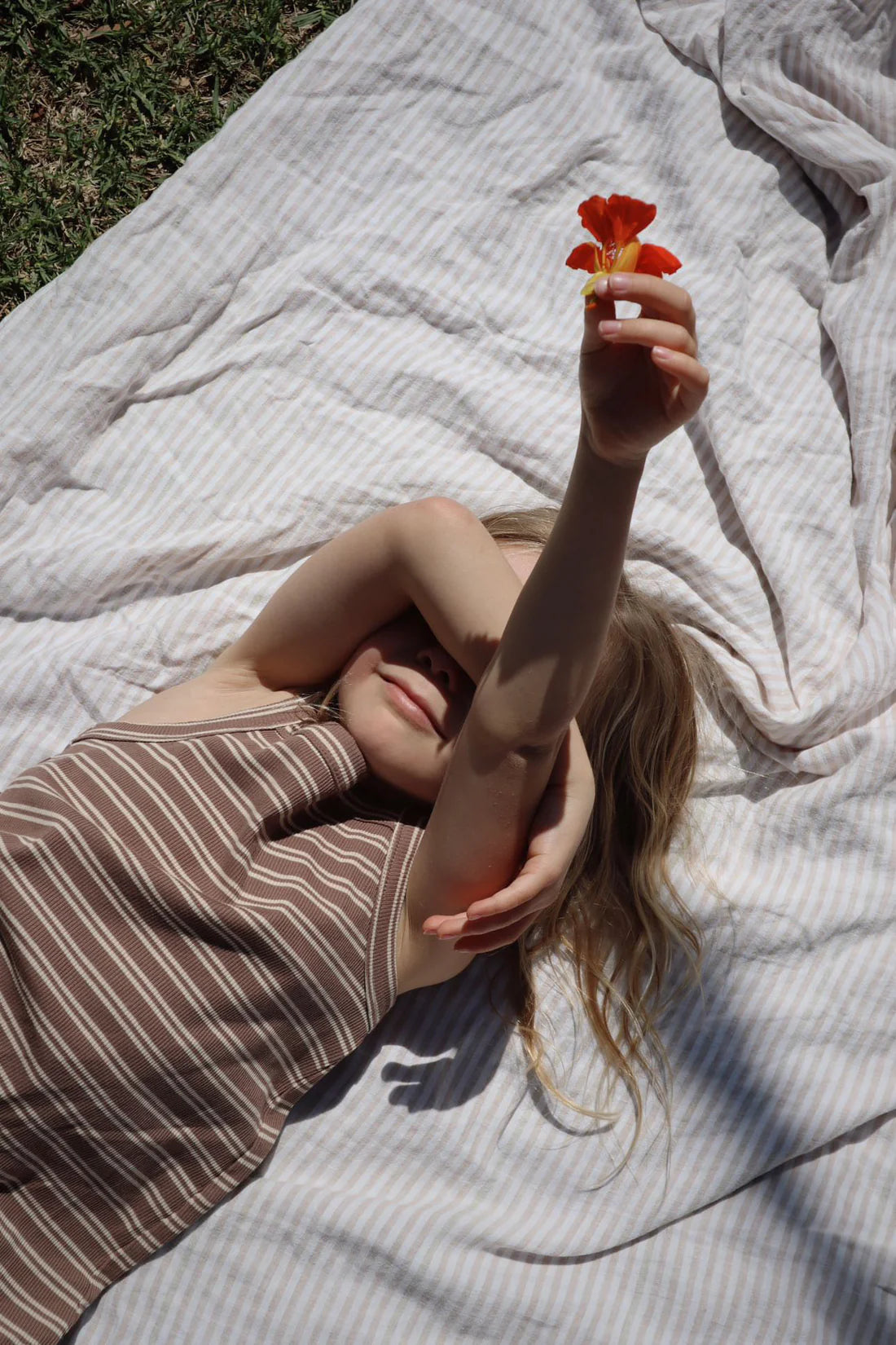 A young girl reclines on a striped blanket outside, smiling with one arm resting on her forehead while the other holds up a small orange flower. She is wearing the Bobbi Rib Dress Choc Stripe by MILKY DESIGNS, featuring brown and white stripes. Sunlight creates gentle shadows around her, accentuating her happiness.