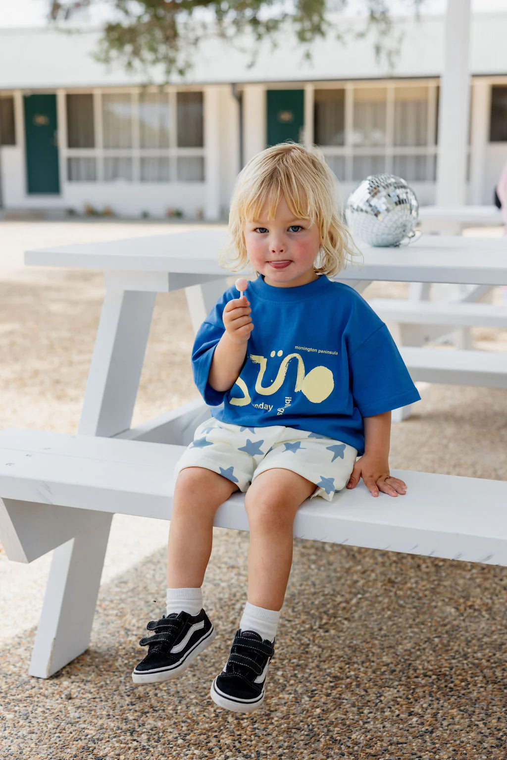 A young child with light blonde hair sits on a white picnic bench, wearing an oversized Il Sole Tee in blue/yellow by SUNDAY SIBLINGS, star-patterned shorts, white socks, and black shoes. A disco ball is beside them as a building stands in the background, adding to the playful scene.