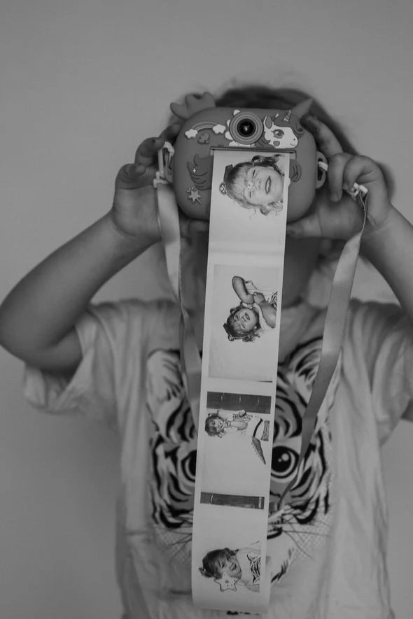 A child showcases creativity with the Oh My Frankie ~ Oh My Snap Instant Print Camera Purple, as they hold it up to their face, a strip of photos capturing playful expressions hanging from it. Their t-shirt features a tiger design. This black and white image captures the joy of discovery.