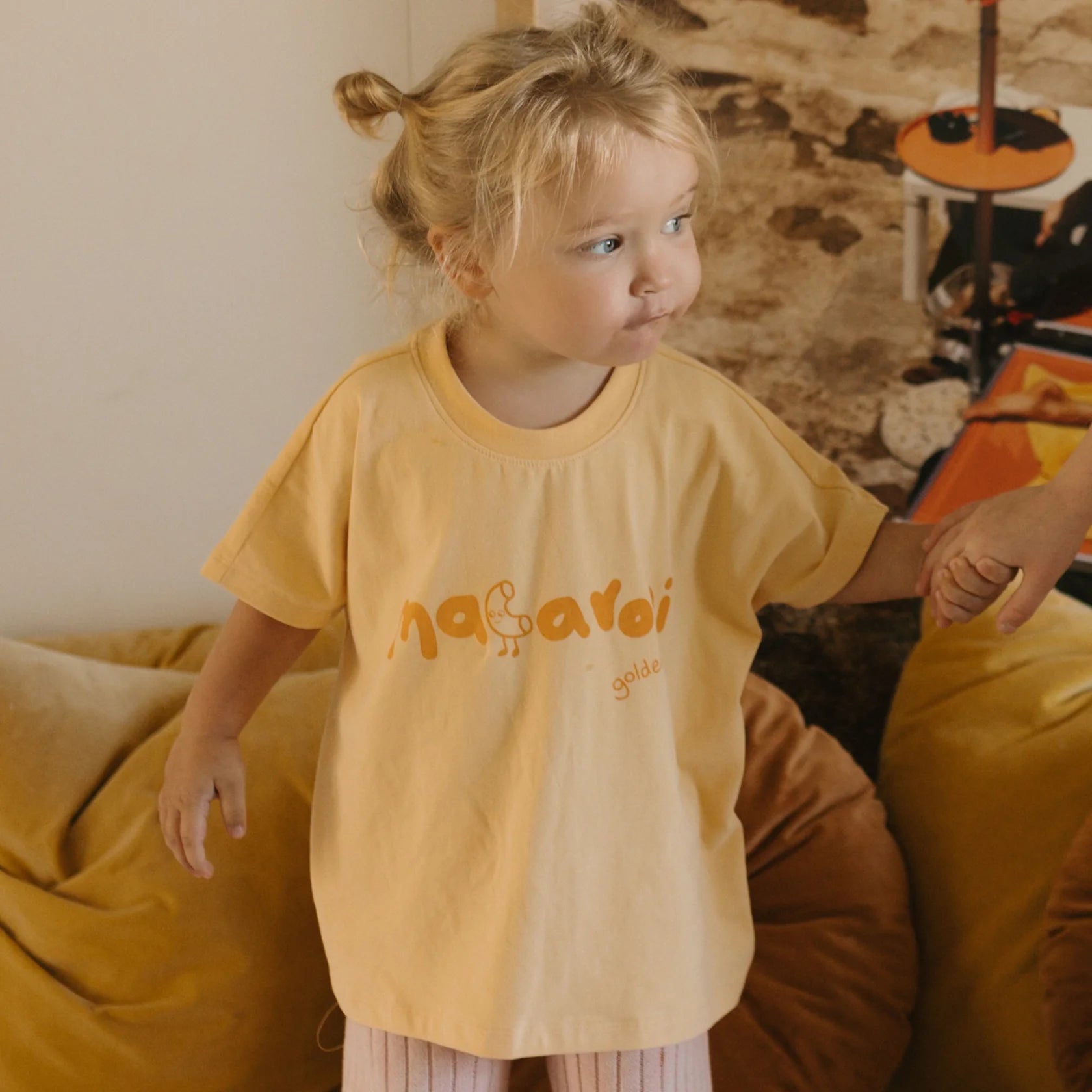 A small child with blond hair is standing and holding an adult's hand. The child is wearing a GOLDEN CHILDREN Macaroni Tee Golden Butter made from soft stretch fabric, featuring the word "macaroni" and an illustration on it. The background includes a cozy setting with light brown cushions and a rustic wall.