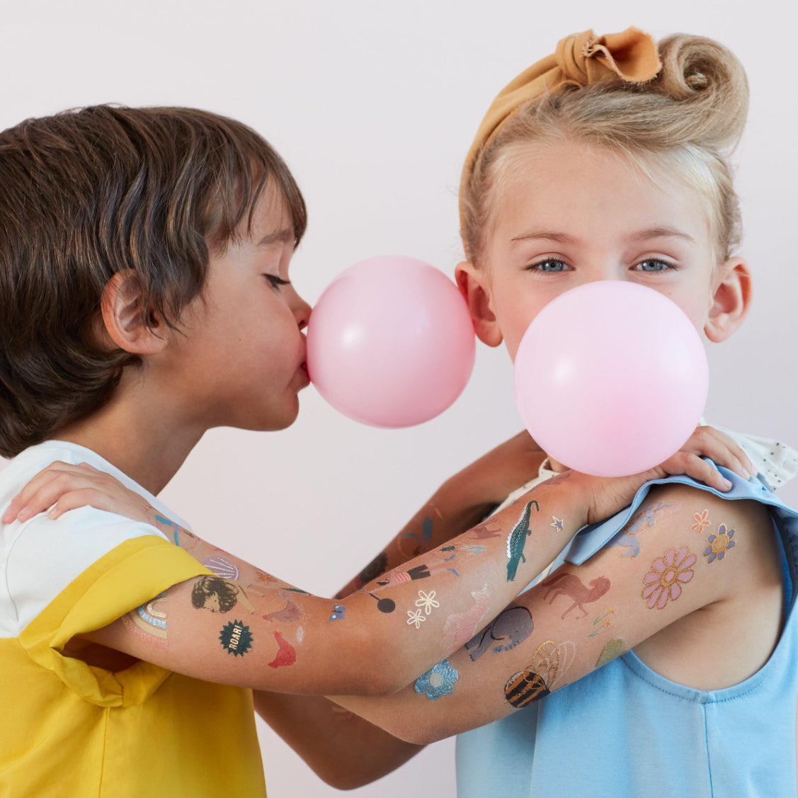 Two young children, adorned with temporary tattoos from the MERI MERI Fairy Tattoos Pack, blow pink bubble gum balloons at a fairy-themed party. The boy sports a yellow and white shirt, while the girl has her hair tied up with a bow and wears a blue top. Both are smiling and having fun.