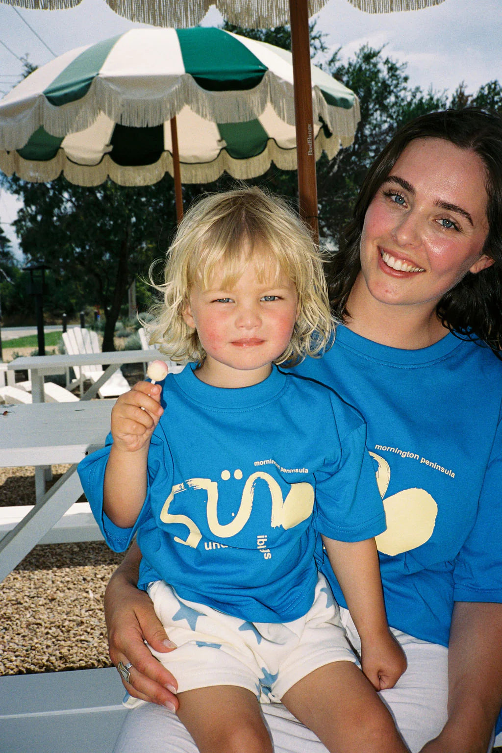 A woman and child, both in matching blue shirts, sit outdoors under an umbrella. The child sits on the woman's lap in adorable SUNDAY SIBLINGS Sunday Siblings ~ Kiddo Shorts Blue, holding a small object and looking at the camera. Picnic tables and trees create a charming backdrop.