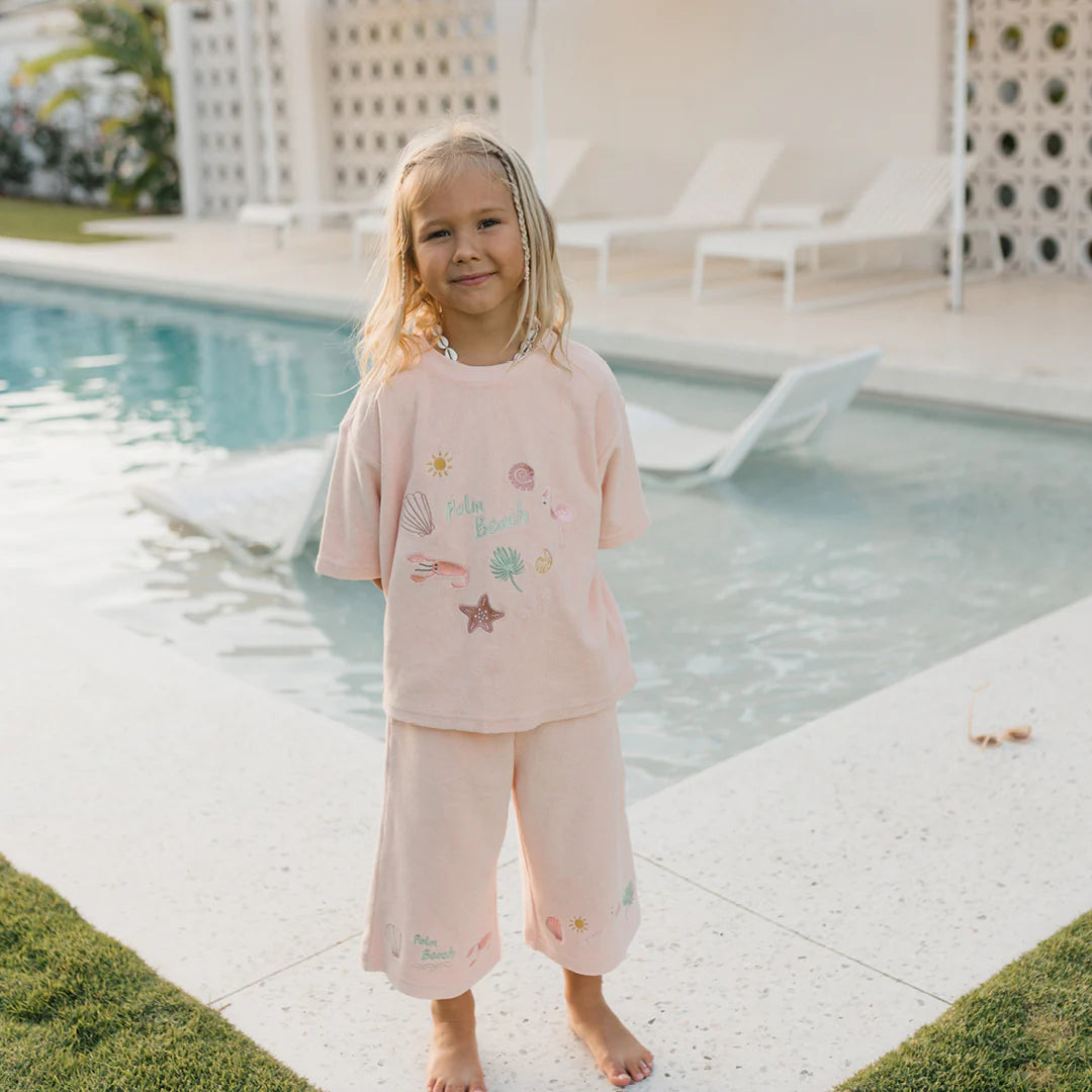 A young child with long hair stands by a pool, smiling at the camera while wearing GOLDEN CHILDREN's Palm Beach Wide Leg Pants in Flamingo Pink, made of terry towel fabric with beach-themed designs. The background shows poolside lounge chairs and a modern outdoor setting.