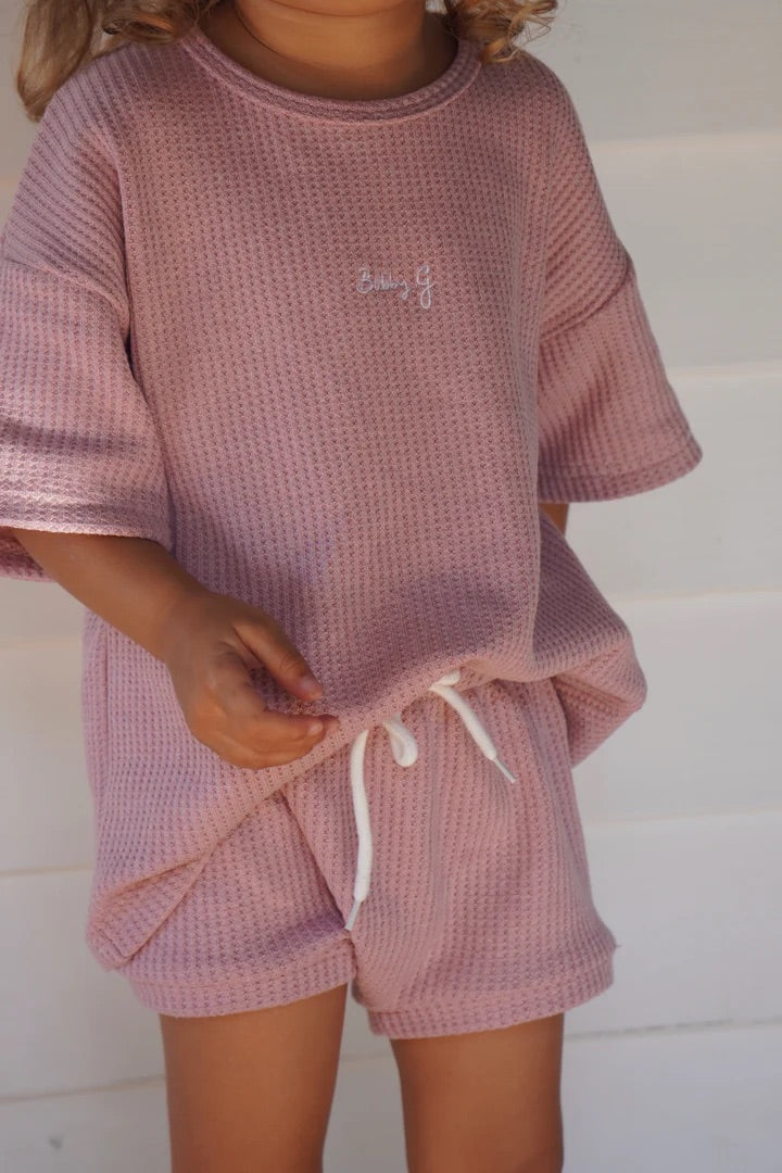 A child wears the Coby Shorties Dusty Rose set by BOBBY G BABY WEAR, featuring a dusty pink waffle-knit top and shorts with a white drawstring. The comfy fabric and stretchy waistband ensure ease, with small chest text, all against a blurred white wall backdrop.