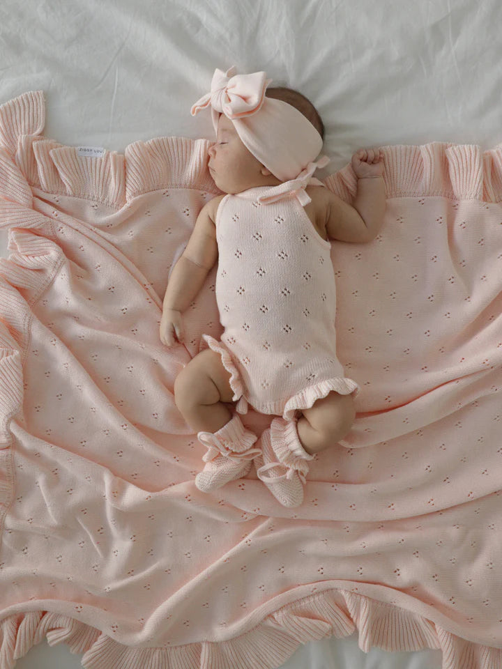 A baby dressed in the Frill Bodysuit Primrose from ZIGGY LOU, complete with a matching headband and booties, sleeps peacefully on a pink blanket adorned with ruffled edges. The 100% cotton outfit and blanket showcase a delicate pattern, achieving a coordinated and serene look as the baby rests on a white surface.