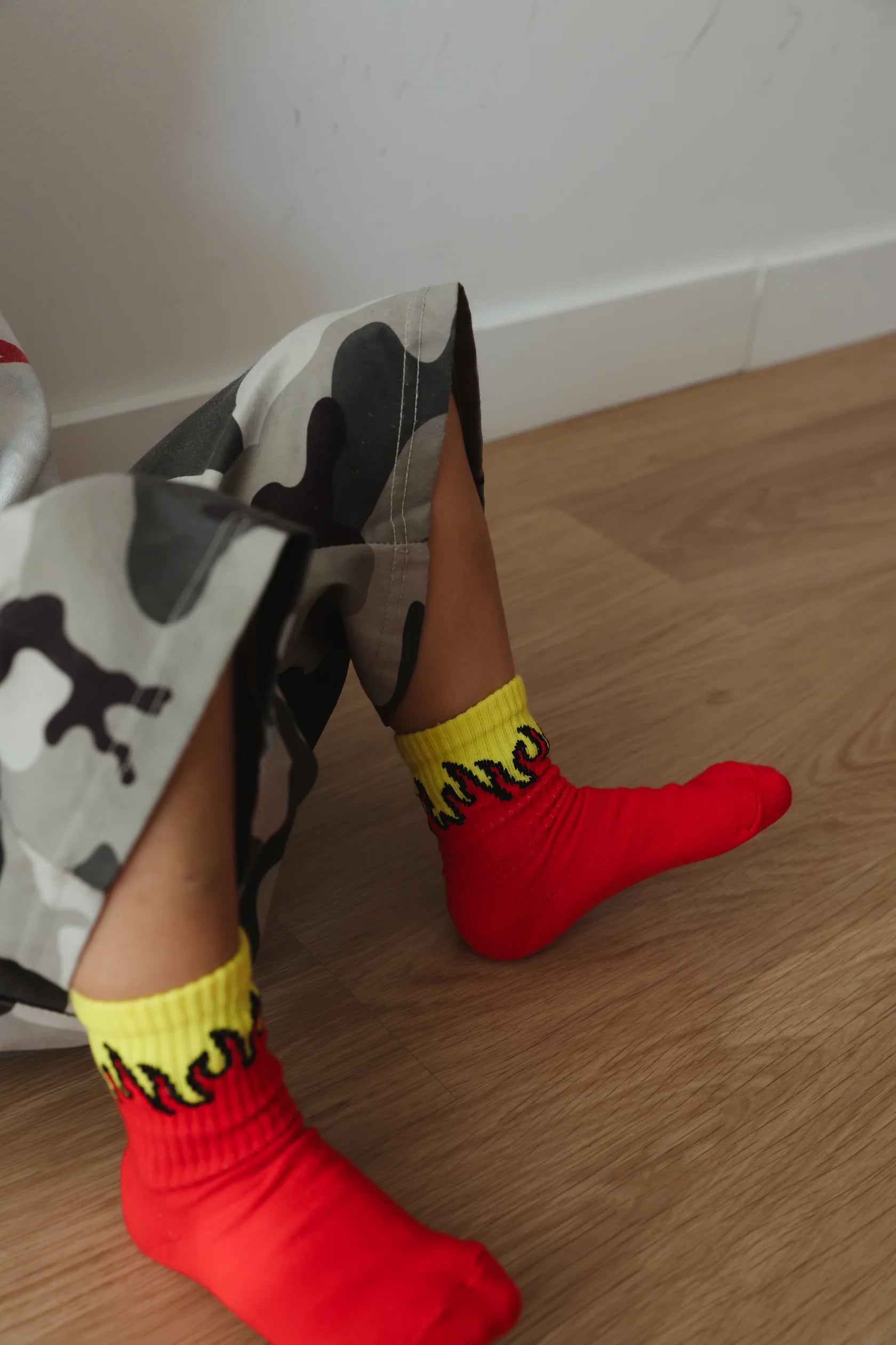 A child wearing camo pants with elastic banded ankles and a pair of TINY LOVE CLUB Fire Socks featuring a bright red color with yellow flame designs sits on a wooden floor.