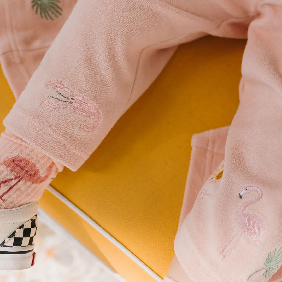 A child dressed in Flamingo Pink Palm Beach Wide Leg Pants by GOLDEN CHILDREN and flamingo socks sits on a yellow surface. The pants, adorned with embroidered animals like a flamingo and lobster, add charm to the scene, while a checkered shoe peeks out playfully.