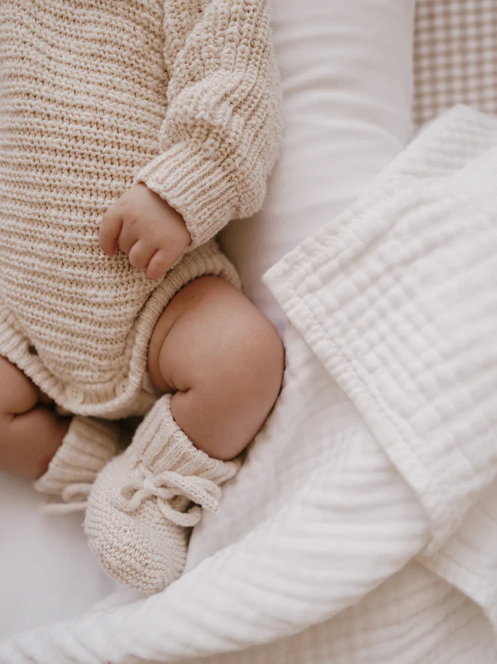 A baby wearing a Heirloom Romper Honey from the ZIGGY LOU collection and matching booties lies on a white quilted blanket. The image focuses on the baby's torso and legs, capturing the texture of the knitted clothing and the soft, cozy environment.