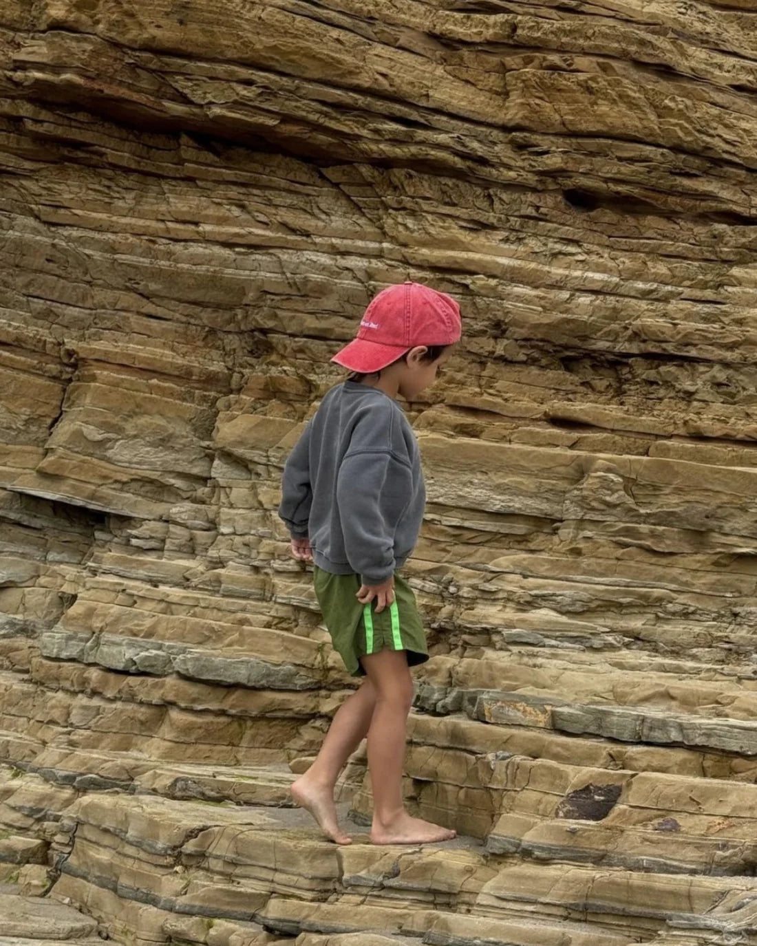 A young child climbs barefoot on rocky terrain, wearing a Ziggy Zaza ~ Roses Are Red cap with an adjustable brass clasp, a gray sweatshirt, and green shorts, appearing focused against the natural stone formations.