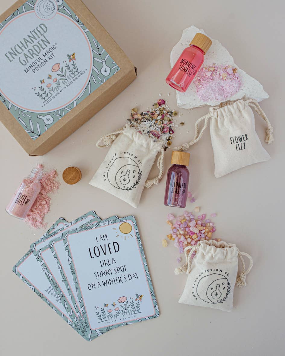 A flat lay image showcases a gift box labeled "Enchanted Garden MINDFUL Potion Kit" by THE LITTLE POTION CO, with small pouches, bottles, and cards arranged around it. The items include "Flower Fizz" bags, small vials with pink and purple liquids reminiscent of a potion-making kit, and cards with inspirational messages.