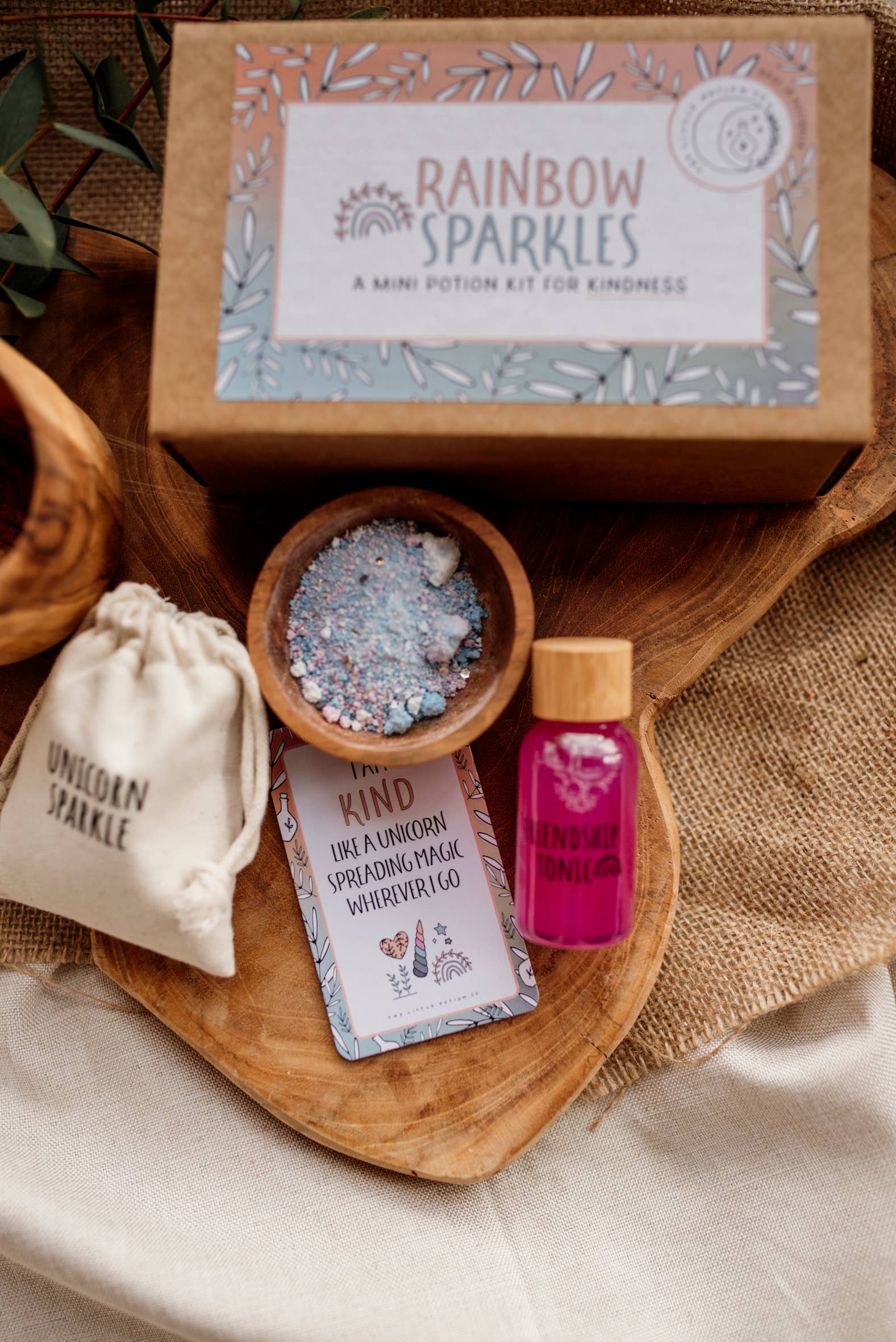 A whimsical flat lay featuring THE LITTLE POTION CO's MINI Rainbow Sparkles Potion Kit (A potion for kindness) designed for imaginative play. The set includes a vial of Friendship Tonic, a bag labeled Unicorn Sparkle, a small wooden bowl with colorful sparkles, and a card inscribed with "Be Kind." All items rest on a wooden surface.