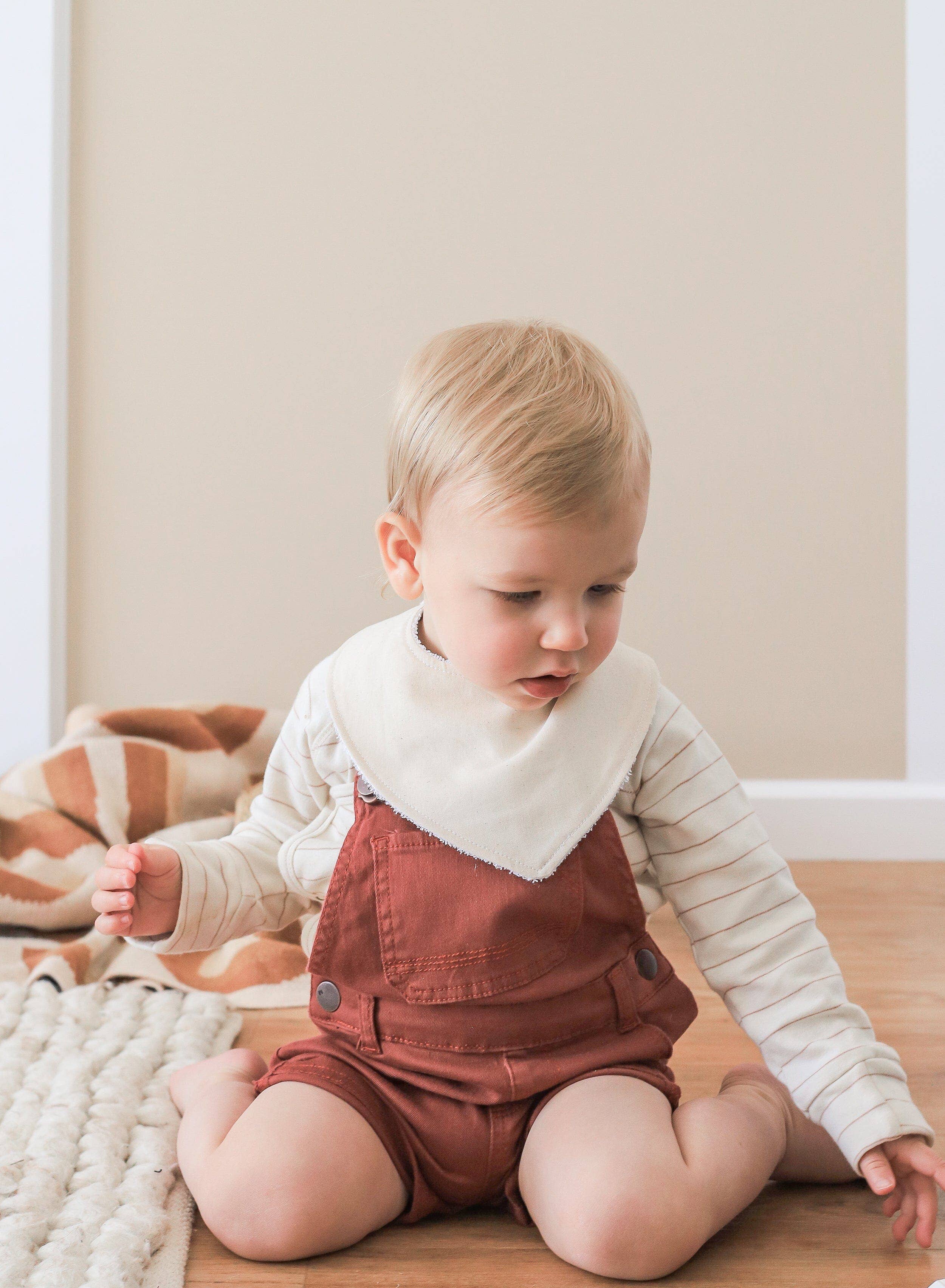 A toddler in rust-colored overalls and a KIIN BABY Dribble Bib Oat with snap fasteners sits on a wooden floor, looking down. A beige and white striped blanket lies nearby against a beige wall.