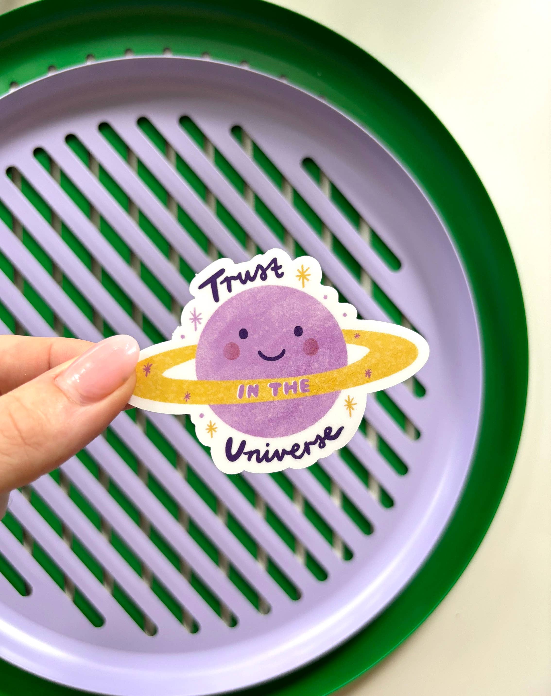 A hand holding a decorative "Trust in the Universe" sticker by LAUREN SISSONS STUDIO, which features a cute, smiling purple planet with a yellow ring and whimsical text. In the background, there is a purple and green circular tray with slotted grooves.