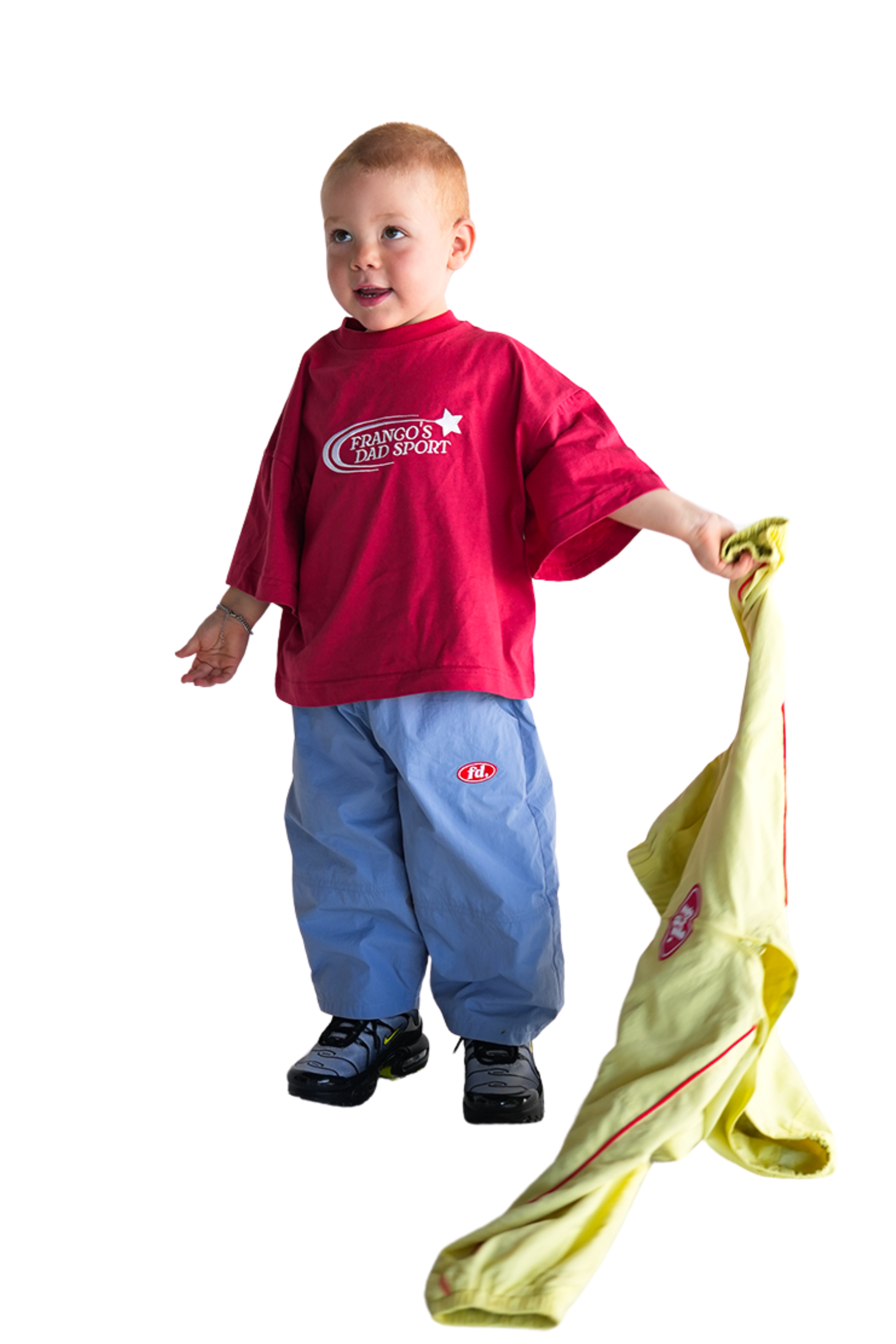 A toddler in Franco's Dad ~ Tech Tracks Blue pants stands on a white background, holding a yellow jacket. They wear a red T-shirt, have short brown hair, and smile with an inquisitive expression. Brand: FRANCO'S DAD.